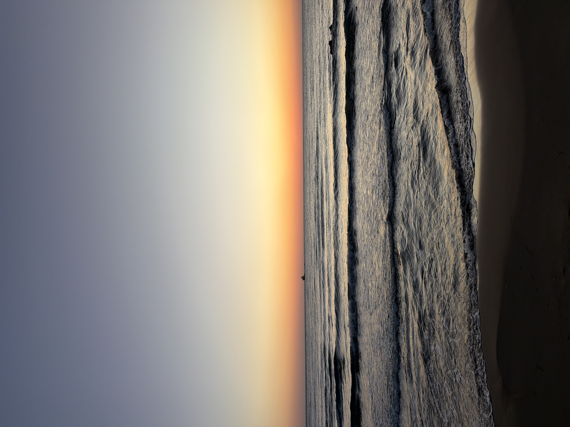 This is a photograph of the sea and beach at Georgioupoli, Greece, during either sunrise or sunset. The sky transitions from a pale blue at the top to a gradient of soft yellow and orange near the horizon. The sea is calm with gentle waves rolling towards the shore. In the distance, a small boat is visible on the water. The foreground shows a dark, sandy beach.