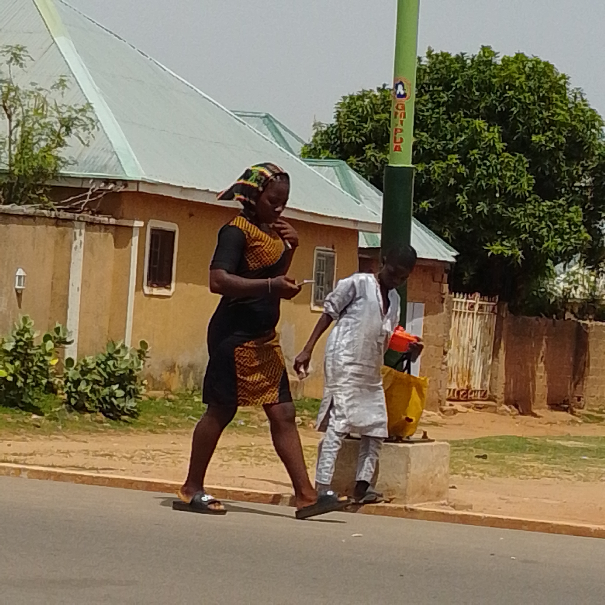 Two individuals walk on a paved road in Gombe, Nigeria. The person on the left is a female wearing a black and yellow patterned dress with a headscarf. She is holding a mobile phone and appears to be looking at it. The person on the right is a male wearing a white traditional outfit and dark sandals. He is carrying a yellow bag and an orange object. They are walking in the same direction, on the right side of the road. In the background, there is a building with a light green roof and beige walls. Trees and a metal gate are also visible. A green pole with markings is positioned between the two individuals and the background. The sky is overcast.