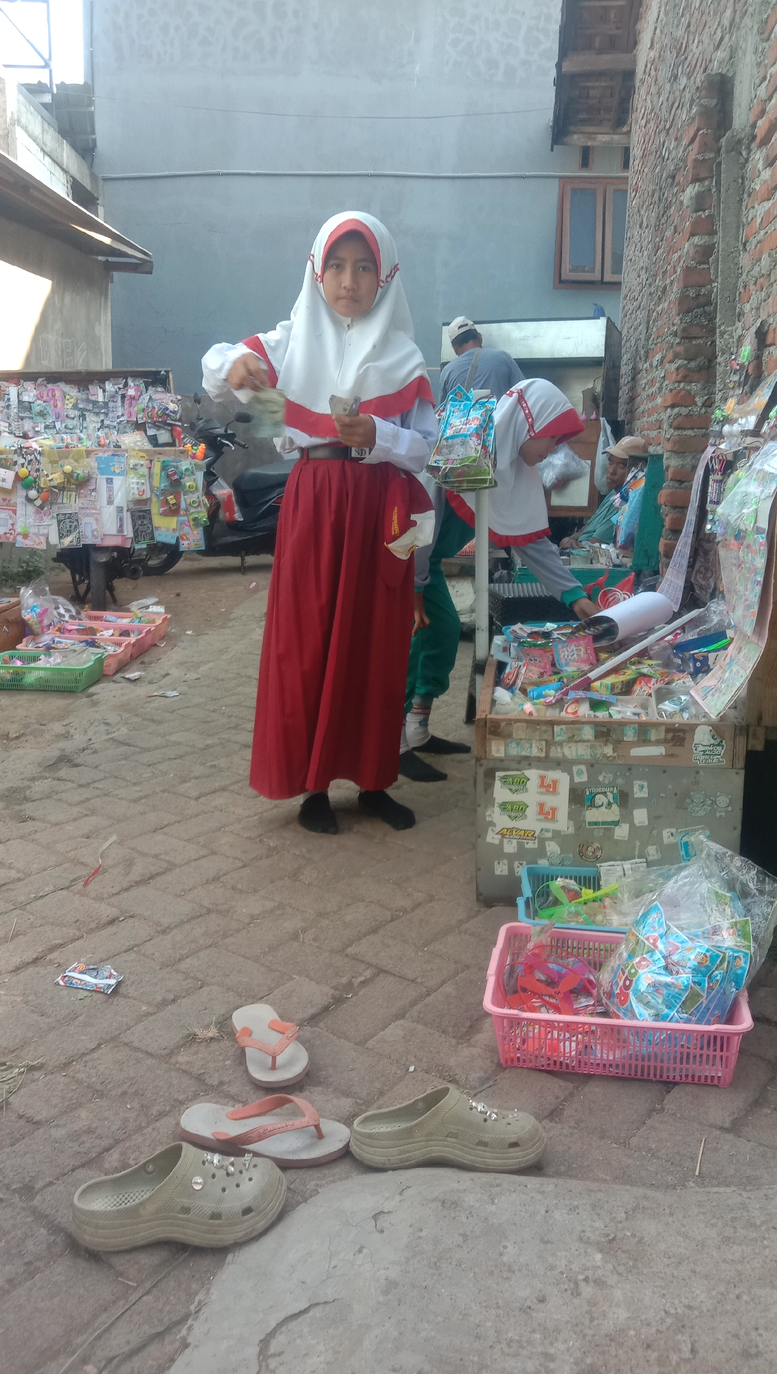 In the city of Karang Anyar, Indonesia, a young girl dressed in a white hijab with red trim and a red skirt stands on a paved walkway. She is holding out a small amount of money in her right hand and appears to be looking at it. Behind her, other children in similar school uniforms are visible, along with a motorbike. To her right is a stall filled with various goods, including packaged candies and toys, some of which are displayed in pink and blue plastic baskets. In the foreground, several pairs of sandals and clogs are scattered on the ground. The scene appears to be outdoors, possibly near a market or street vendor.