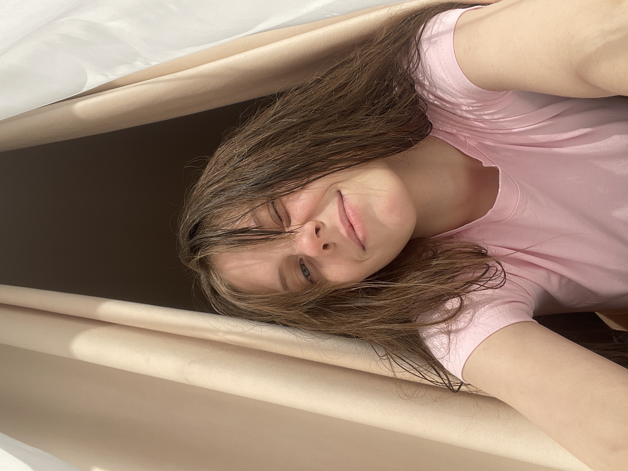 A young woman with wet, dark brown hair is centered in the frame, wearing a light pink T-shirt. She has pale skin and blue eyes, and she is looking at the camera with a slight smile. The background consists of beige fabric that appears to be part of a draped covering or tent. The lighting suggests an indoor setting. There is no indication of the city of Ufa, Russia, in the image. The scene appears to be a casual, close-up self-portrait.