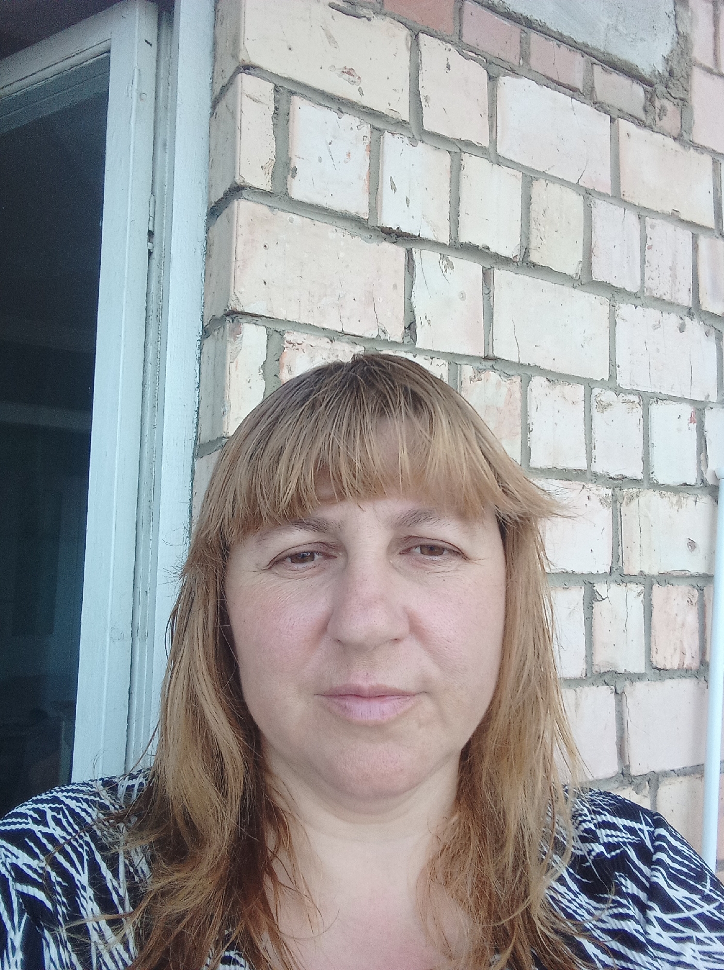A medium-shot, eye-level, self-portrait photograph captures a woman with shoulder-length blonde hair and bangs standing in front of a light-colored brick wall. She is wearing a dark blue and white patterned top. To her left, a portion of a light-colored door frame is visible. The scene appears to be outdoors, possibly a residential area, in Chernivtsi, Ukraine.