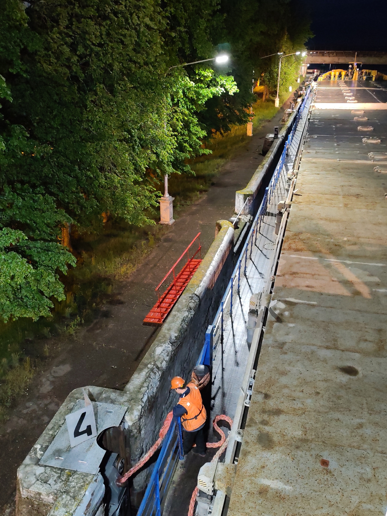 The image shows a segment of the lock in Selivanovo, Russia. A worker wearing a safety vest and helmet stands behind a blue railing, holding a rope connected to a mechanism. The lock wall features a sign with the number "4". A red ladder is visible leading down from the lock wall to the adjacent ground area, where trees and streetlights are present. The lock extends into the distance under the illumination of artificial lights.
