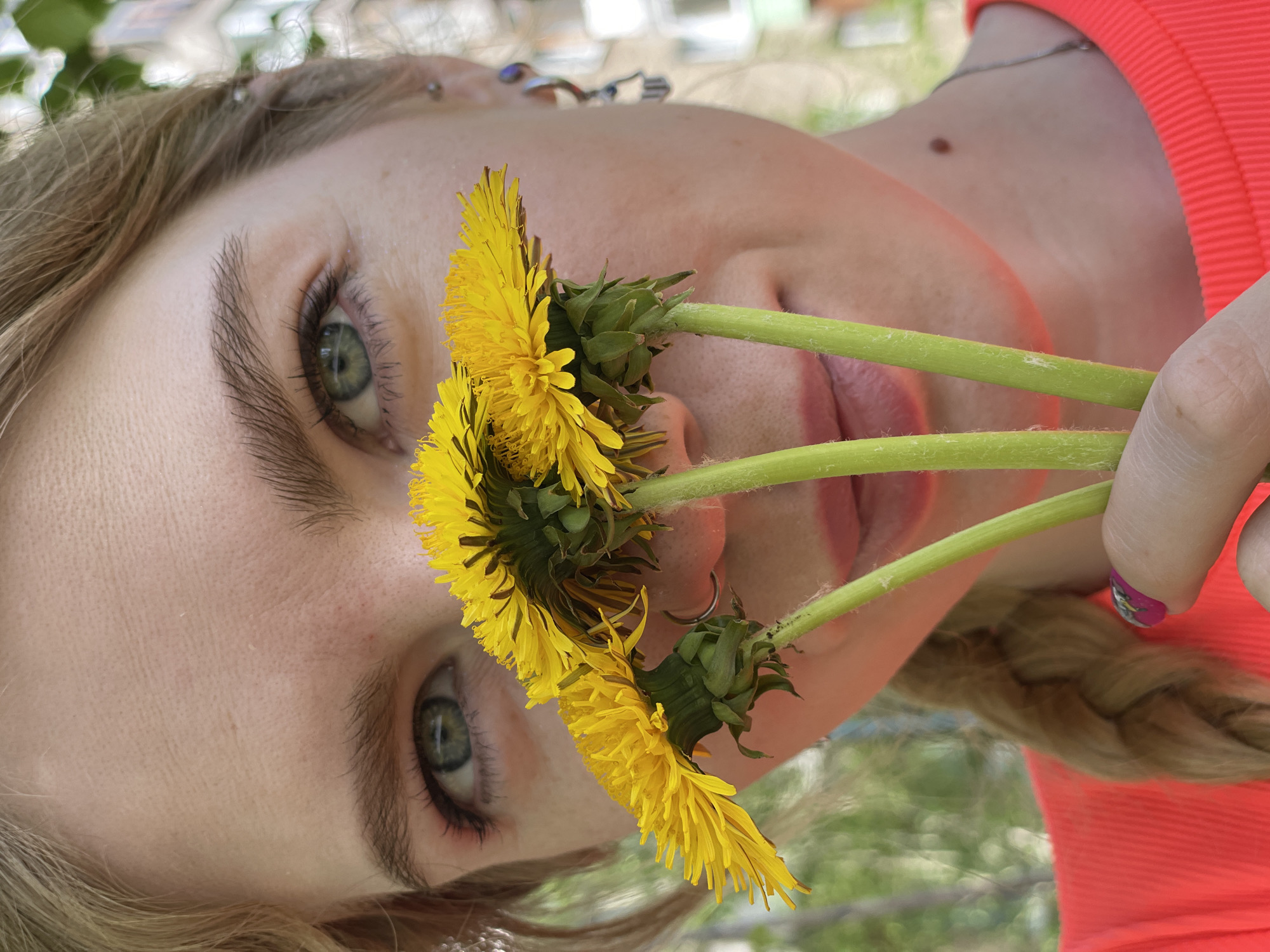 A close-up, rotated image features a fair-skinned young woman holding three dandelions in front of her face, obscuring her nose and part of her mouth. She has green eyes with brown eyelashes and eyebrows. Her blonde hair is braided and visible near her shoulder. She wears a bright pink or coral-colored ribbed shirt. A small mole is visible on her neck. Her fingernails are manicured with a pink and purple design. The dandelions have vibrant yellow petals and green stems. The background is blurred and appears to be outdoors, with indistinct greenery and what might be buildings. There is no specific information in the image that confirms the location as Izhevsk, Russia.
