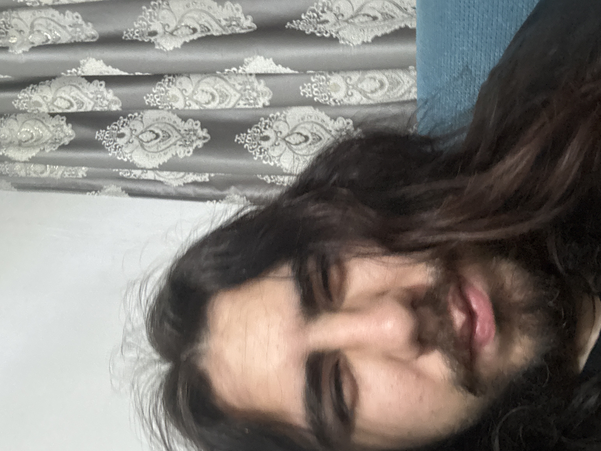 The media file is a close-up, self-portrait-style photograph of a person with long, dark hair. The person's face is angled towards the camera, with their eyes partially closed and their mouth slightly open. The image is heavily cropped, focusing on the individual's features. The background consists of a patterned gray curtain with white paisley designs and a blurred blue textured surface, possibly a piece of furniture. The lighting appears to be indoor, with no clear indication of the time of day or weather. The overall impression is casual and intimate, possibly taken in a home setting. There is no visible text in the image.