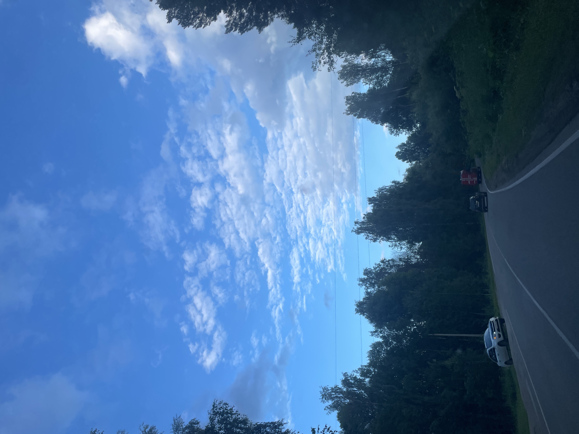 The image depicts a roadside scene under a bright blue sky with scattered white clouds. The perspective is from inside a vehicle, looking out at a winding asphalt road bordered by dense green trees and foliage. Two vehicles are visible on the road: a red truck and a white SUV. The white SUV is parked on the right shoulder of the road, facing away from the viewer. The red truck is further ahead and also appears to be parked or moving slowly. Overhead, power lines are visible stretching across the sky, cutting through the trees. The lighting suggests it is daytime, likely late afternoon or early evening, given the shadows cast by the trees. The overall mood is serene, with the natural landscape dominating the scene. There is no visible text in the image.