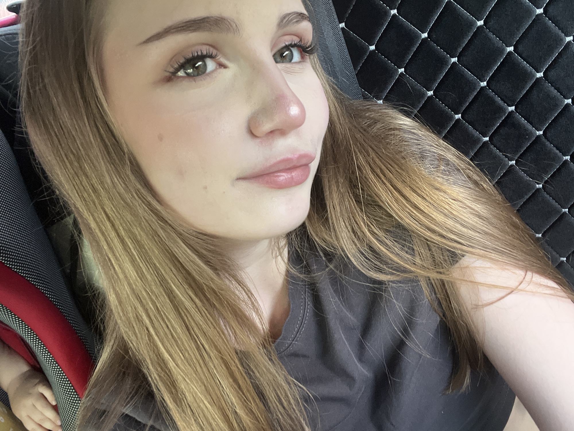 A close-up, low-angle, over-the-shoulder selfie features a young woman with long, blonde hair, a grey t-shirt, and a partially visible baby hand in the upper left corner. She is lying down on a quilted black surface, possibly a car seat or stroller liner. The background is blurred, with hints of red and white fabric. The lighting is soft and diffused, with a focus on the woman's face, highlighting her light green eyes, pink lips, and fair skin. Her hair is styled naturally, with some strands falling across her face and neck. The image appears to have been taken indoors, possibly in a vehicle. There is no information available about the location in the city of Загородный, Russia.