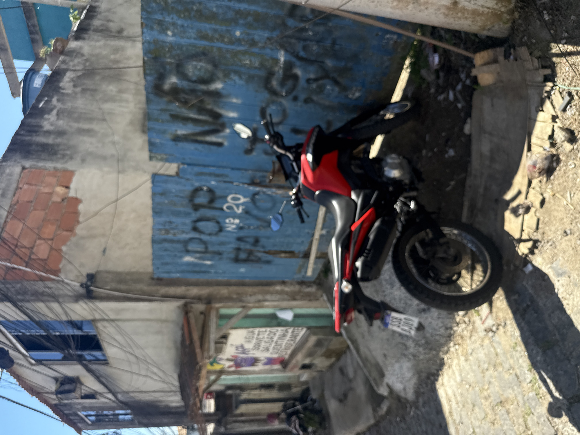 The image depicts a narrow street in Arraial do Cabo, Brazil, likely a residential or commercial area within a favela. The scene is characterized by a mix of weathered buildings, exposed electrical wiring, and a blue corrugated metal fence with graffiti.

In the foreground, a red and black motorcycle is parked on a dirt and stone-covered ground. The motorcycle's license plate is partially visible, reading "SSB 6369". Behind the motorcycle, the blue fence features spray-painted text, including "POP" and "NAO" which likely form part of a larger message.

To the left of the motorcycle, a sign is attached to a wooden structure, appearing to advertise "AGUA" (water), "SORVETE" (ice cream), and "QUENTINHA" (hot meal), suggesting a small vendor or food establishment. Further down the street, out of focus, a person is partially visible, seated near some bags, adding a human element to the scene.

The buildings in the background show signs of age and use, with exposed brickwork, cracked plaster walls, and multiple windows. Overhead, a dense network of electrical wires suggests the informal and often unregulated nature of infrastructure in such areas. The overall environment is sunny, indicating daylight hours.