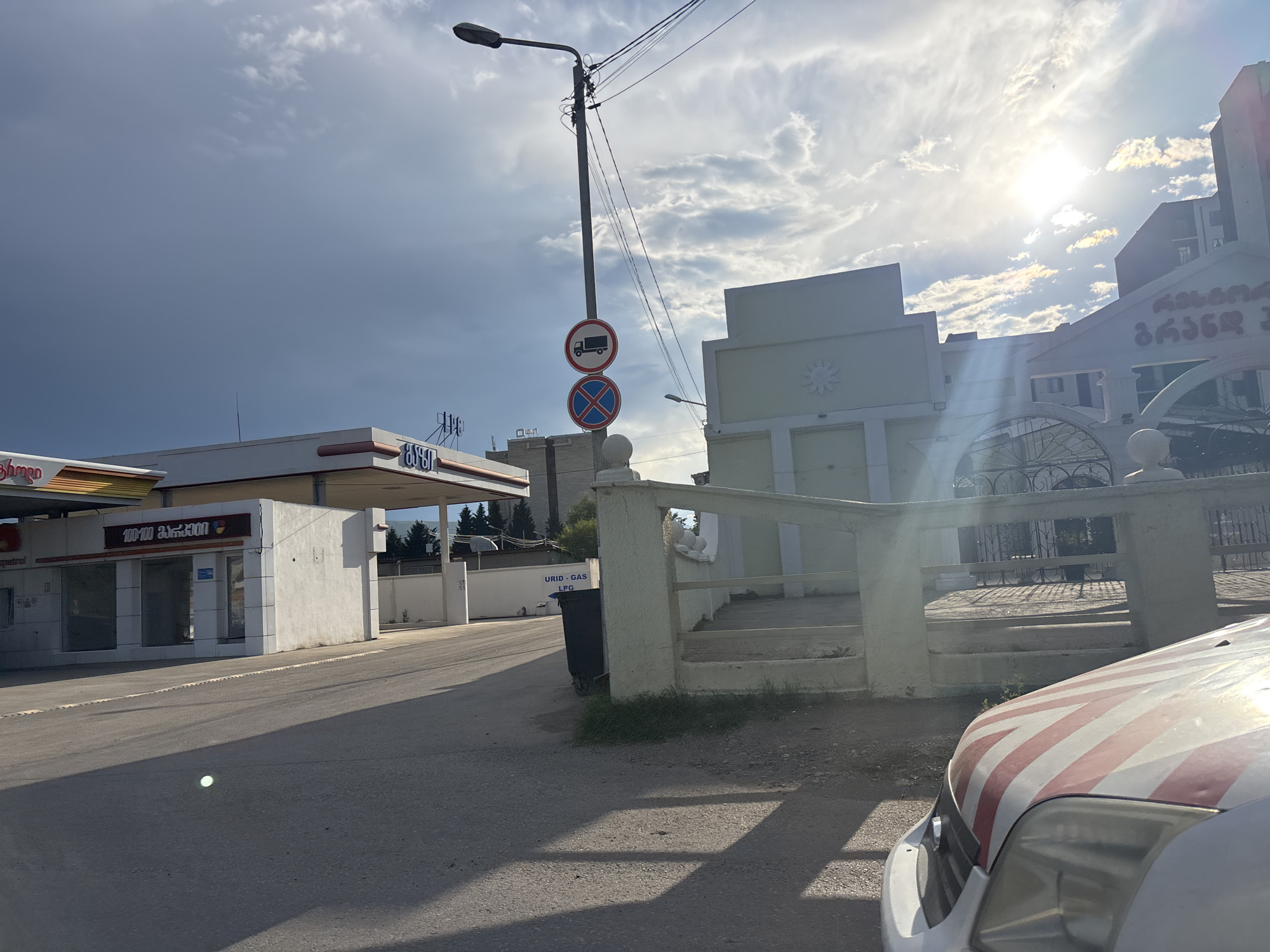 The image depicts an outdoor scene on a sunny day with a mix of clouds. The foreground shows the hood of a car with red and white diagonal stripes. Behind the car is a paved area and a sidewalk.
In the middle ground, a modern building with white and light green facades stands. To the left, a gas station structure with a white roof and columns is visible. The gas station appears to be operational, with signage and pumps. There are also traffic signs posted on a lamppost, including a "no trucks" sign and a "no stopping" sign. Further back, another building with an arched entryway and decorative elements is seen. A black trash bin is placed near a low wall.
The background is dominated by a bright sky with sunbeams breaking through scattered clouds. The setting appears to be urban or suburban, possibly on the outskirts of Tbilisi, Georgia, given the text visible. The text "URID - GAS LPG" is written on a white sign on a wall behind the gas station. The Georgian text "100-100 გორგოვა" is visible on the side of the gas station building. Another building in the distance has Georgian text that appears to read "ქართული ღვინო" (Georgian Wine). The time of day seems to be late afternoon, indicated by the angle of the sunlight and the shadows. The overall mood is calm and ordinary, capturing a slice of everyday life.