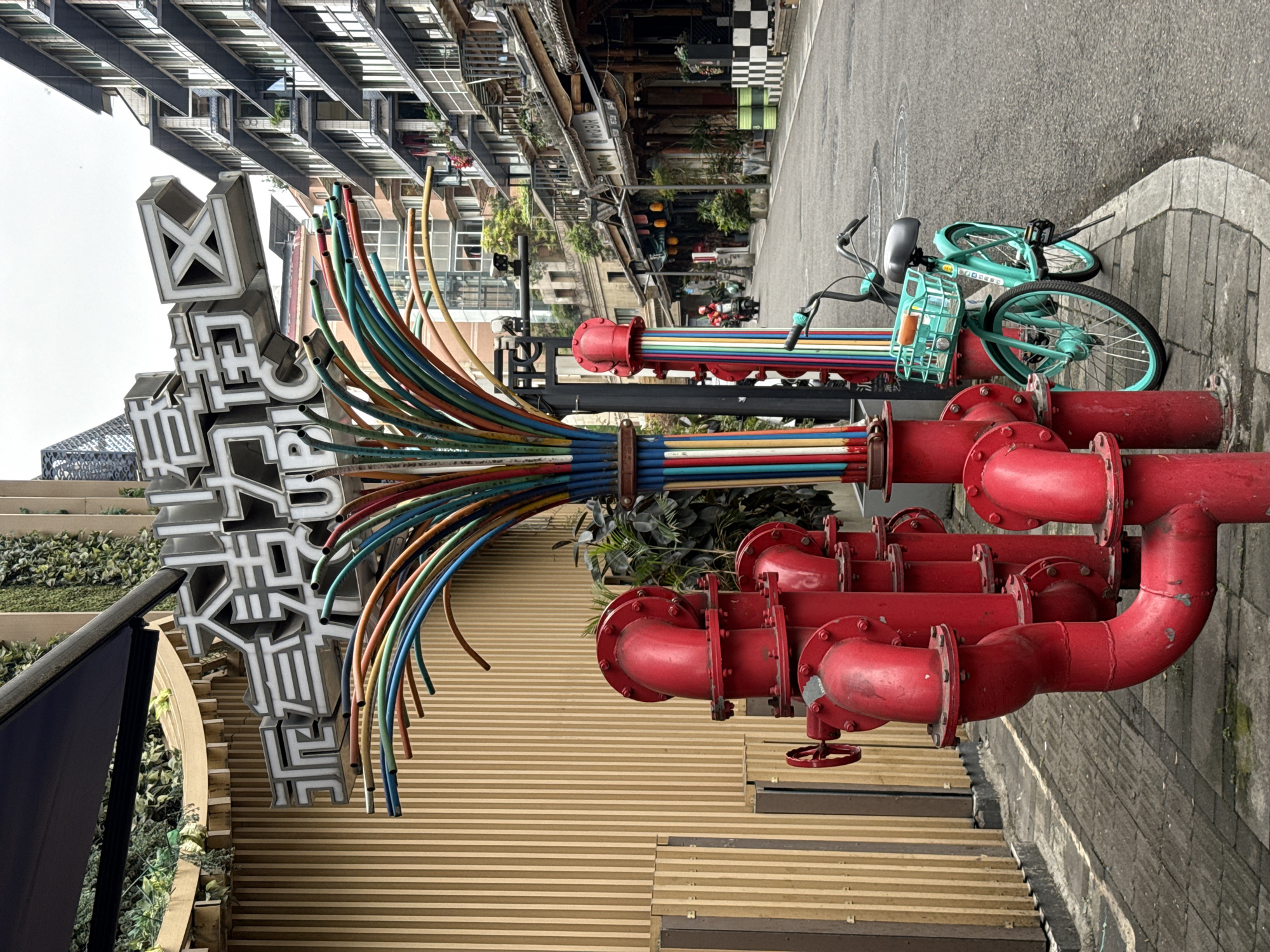 The image depicts a street scene in Jinjiang District, China, featuring colorful urban art installations. One installation consists of numerous brightly colored tubes emerging from a gray column with Chinese characters on top. Another installation involves a series of red pipes with flanges and fittings. A teal bicycle rests near the pipe sculpture. The background reveals urban structures, including buildings with balconies and awnings. The street is paved with gray stone. The overall scene suggests a vibrant, artistic urban environment.