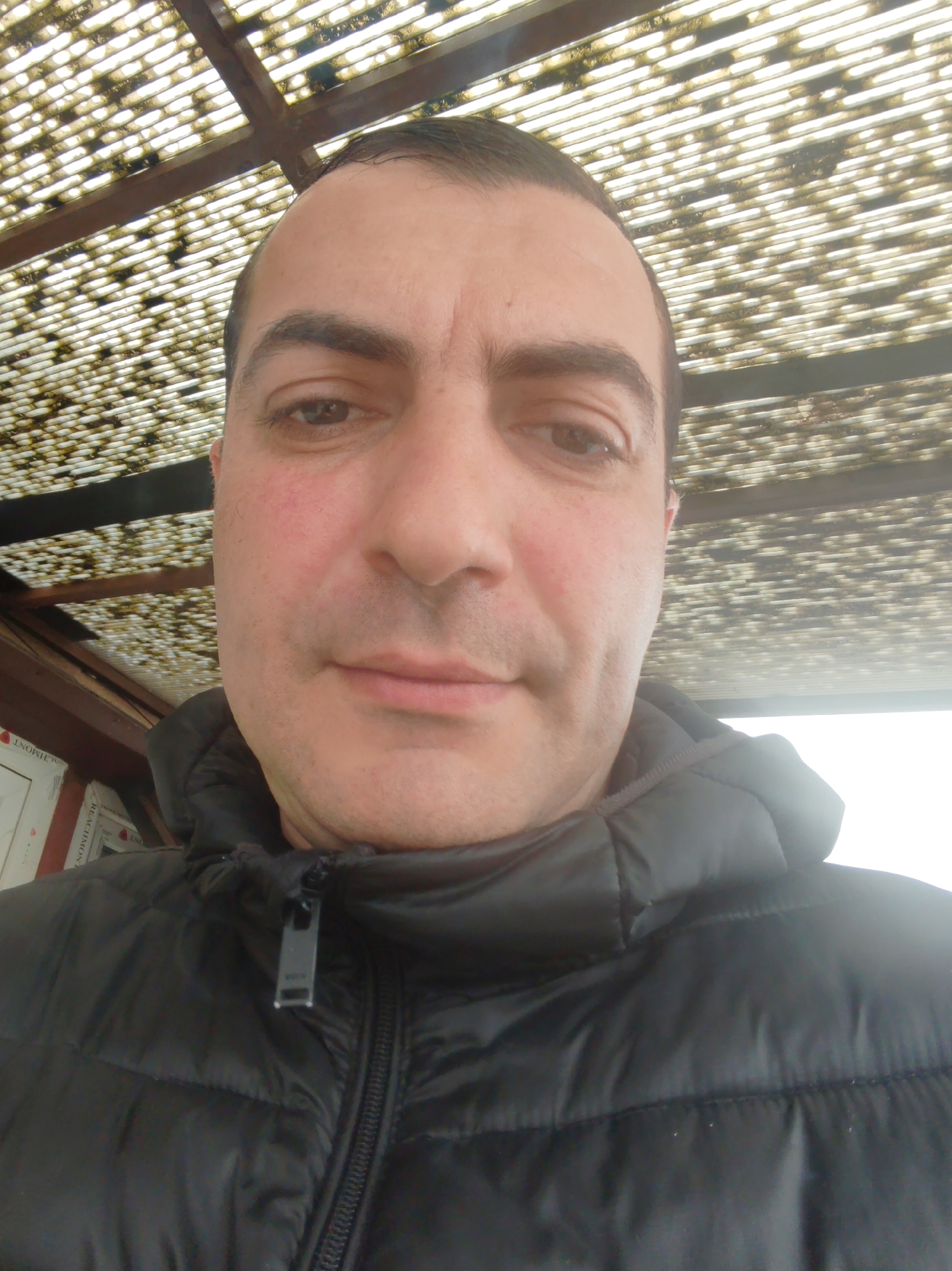 A close-up selfie shows a middle-aged man with dark hair and eyes. He has fair skin with some redness on his cheeks. He is wearing a black puffer jacket with the zipper partly zipped. The background shows a wooden and plastic corrugated roof structure. The location is in the city of Unknown, Unknown.