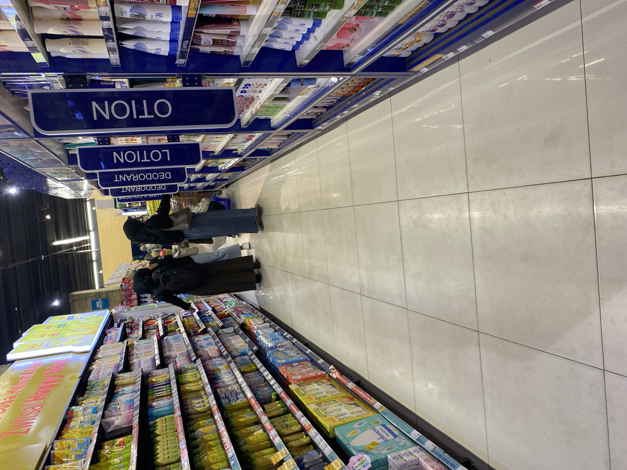 The image depicts a wide aisle in a supermarket in Banda Aceh, Indonesia, filled with rows of shelves stocked with various personal care products, primarily lotions and deodorants. Two women are visible in the aisle. One, wearing a dark hijab and an earth-toned abaya, is reaching towards a shelf on the left. The other woman, also in a hijab and jeans, is standing further down the aisle on the right, looking at products on a shelf. The shelves on the right are organized by product type, with prominent blue signs indicating "LOTION" and "DEODORANT." The floor is tiled with a light-colored material, and overhead lighting illuminates the scene. The overall impression is that of a well-stocked retail environment for personal hygiene items.