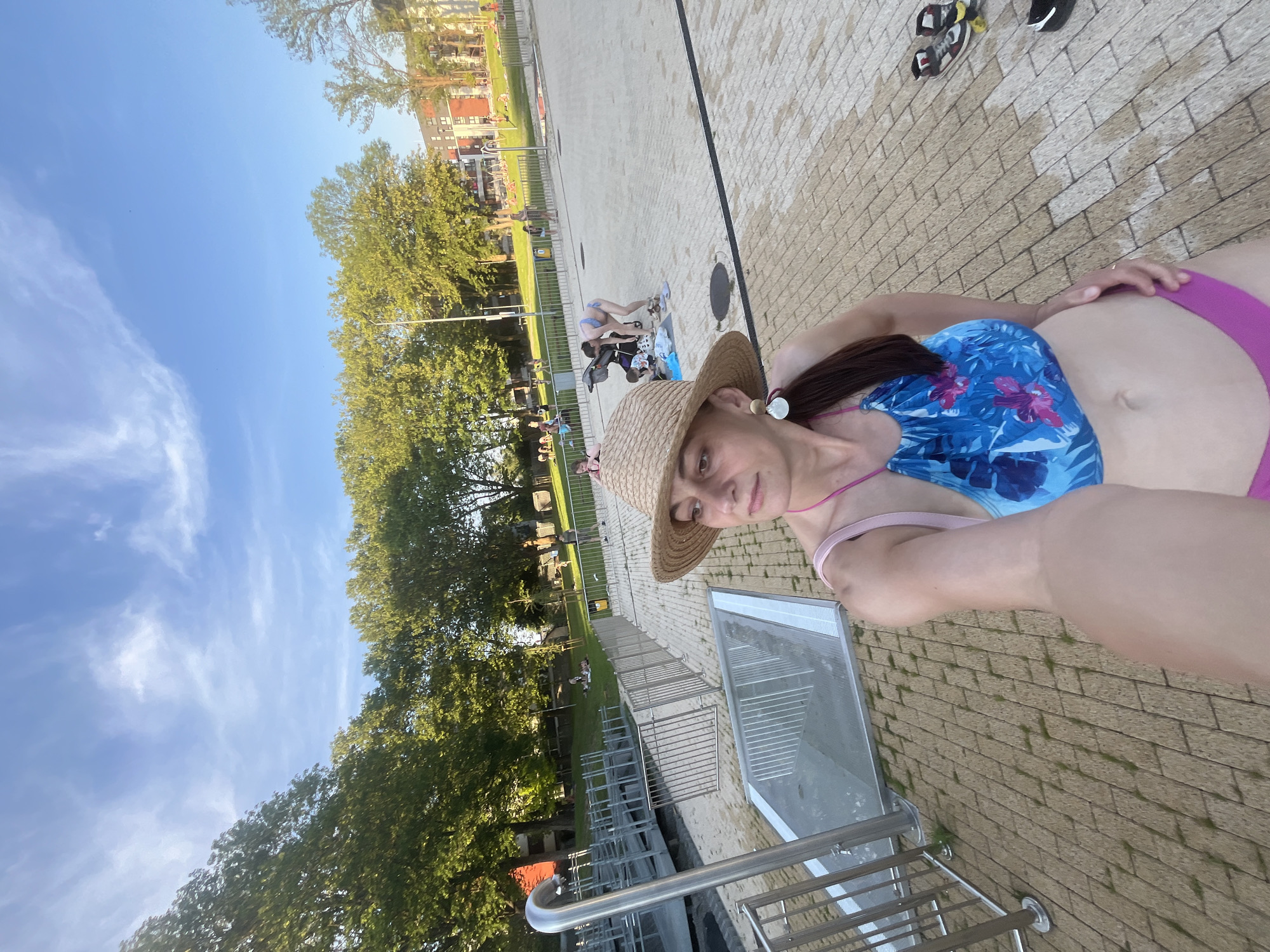A woman in a blue and pink floral bikini and a straw hat takes a selfie outdoors. She has brown hair pulled into a ponytail and is wearing large circular earrings. Behind her, a paved area leads to a grassy park with trees and buildings. Several other people are visible in the background, some sitting on the grass and others near a playground. The sky is blue with scattered white clouds.