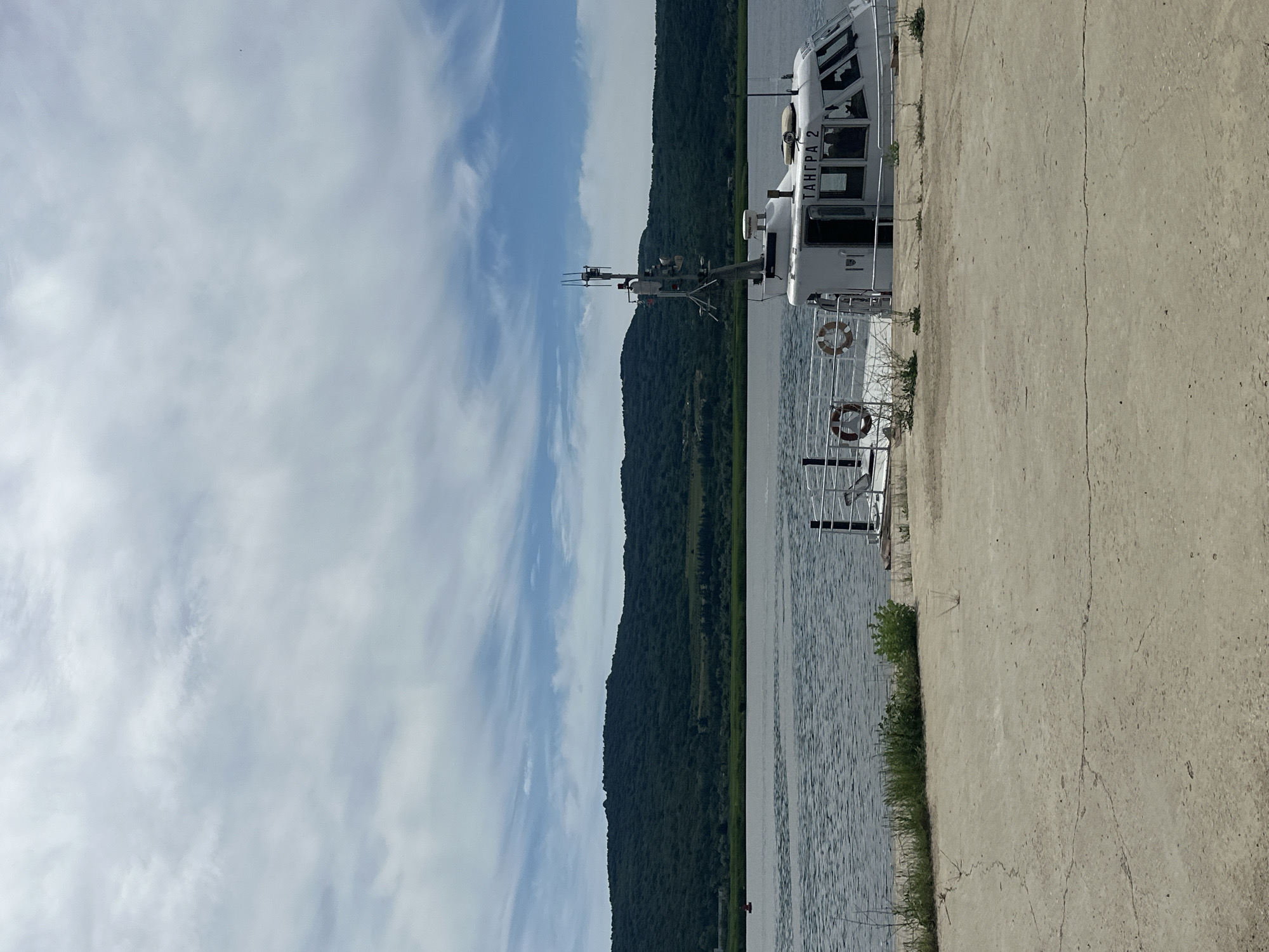 The image depicts a white boat docked on a concrete pier next to a body of water. The boat, labeled "ТАНГРА 2" in Cyrillic script, has a cabin with windows and a radar antenna on its roof. Two life preservers are attached to the railing of the boat. In the background, a lush green, tree-covered hill rises from the opposite shore, under a sky filled with white, wispy clouds. The concrete pier is cracked and weathered, with sparse green vegetation growing in patches. The water appears calm, with ripples on its surface. The scene is set in an outdoor environment, likely a waterfront area.
