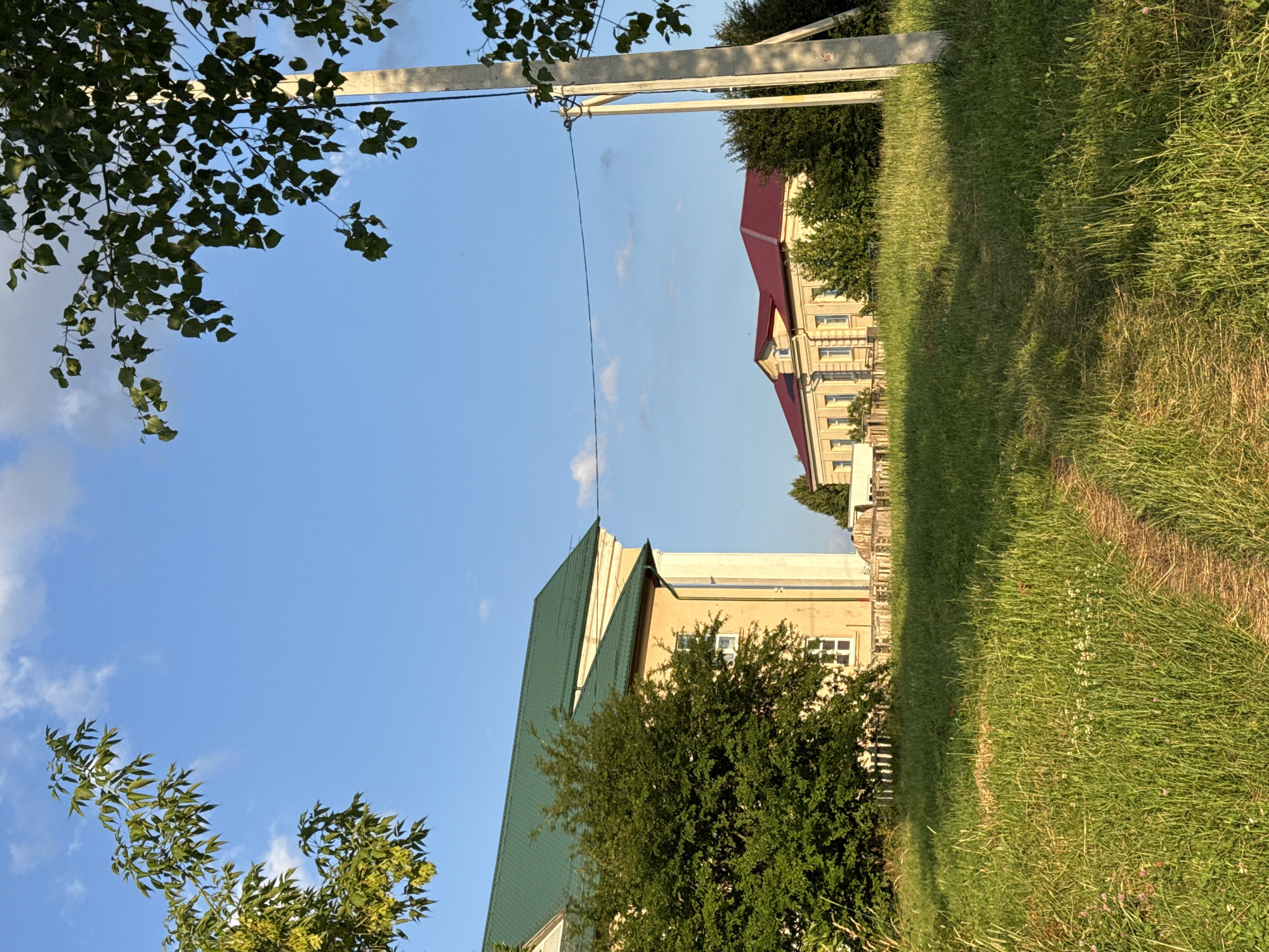 The image depicts a sun-drenched, rural landscape in Zhodzina, Belarus. In the foreground, a dirt path winds through tall, green grass dotted with small white and pink wildflowers. To the left, a large building with a green, pitched roof and light-colored walls with white window frames is partially obscured by a dense, green bush. Further in the distance, a second, long building with a dark red, pitched roof and similar light-colored walls with multiple windows is visible, surrounded by trees. A concrete utility pole with electrical wires stretching across the frame stands to the right of the path, casting a shadow on the grass. The sky is a clear blue with a few scattered white clouds. The scene conveys a sense of peaceful, rural living.