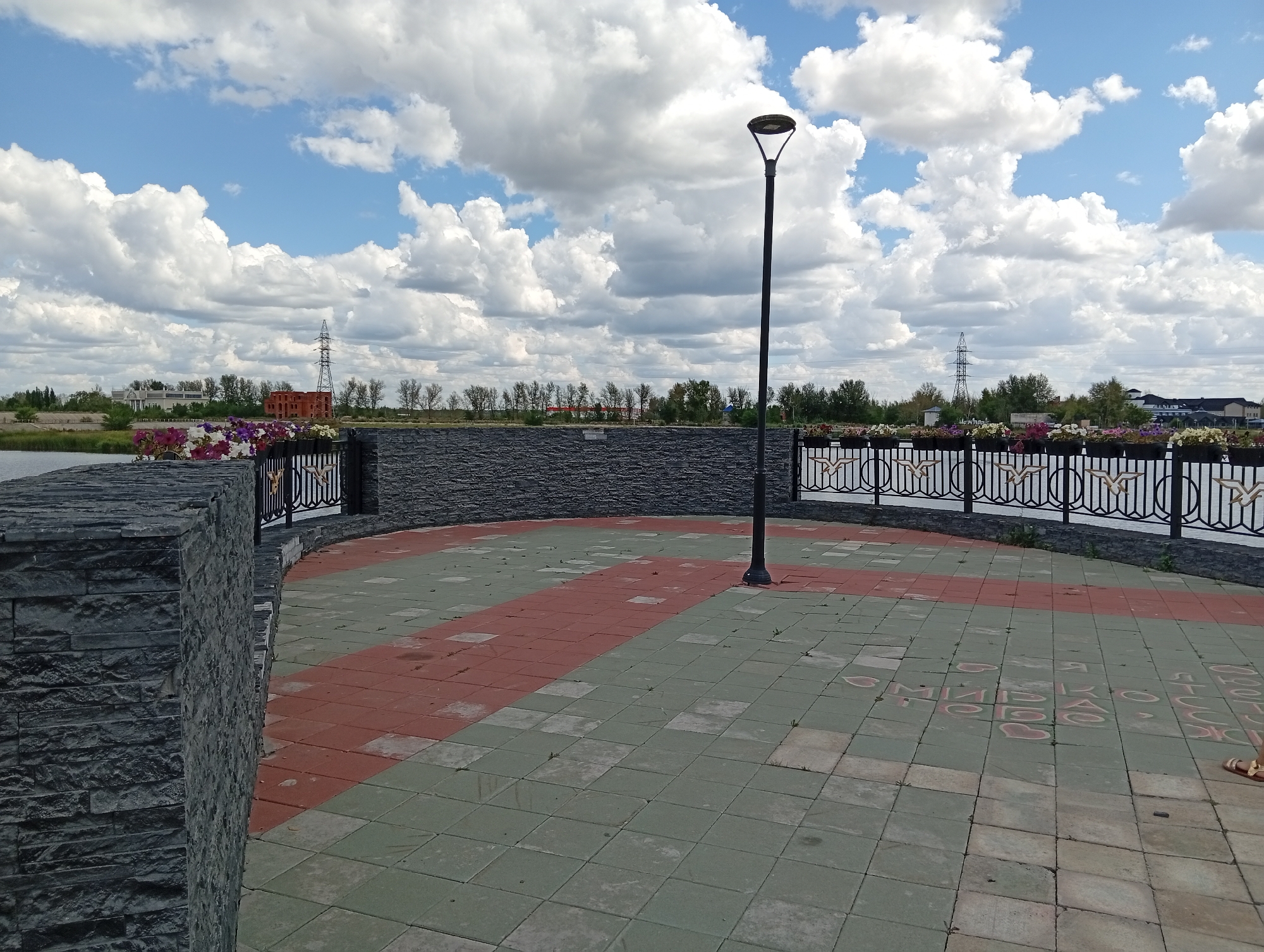 The image shows an outdoor paved area with a street lamp in the center. The paving stones are arranged in a diagonal pattern of red and green squares. A black metal fence with decorative elements runs along the edge of the paved area, separating it from a body of water. Flower boxes filled with colorful flowers are placed along the fence. In the background, there are trees, some buildings, and power lines, under a sky with fluffy white clouds. The scene appears to be a public space, possibly a waterfront promenade or park. Based on the provided location of Заречный ауылдық округі, Kazakhstan, this image depicts a scene within that rural district. There are no people visible in the image.