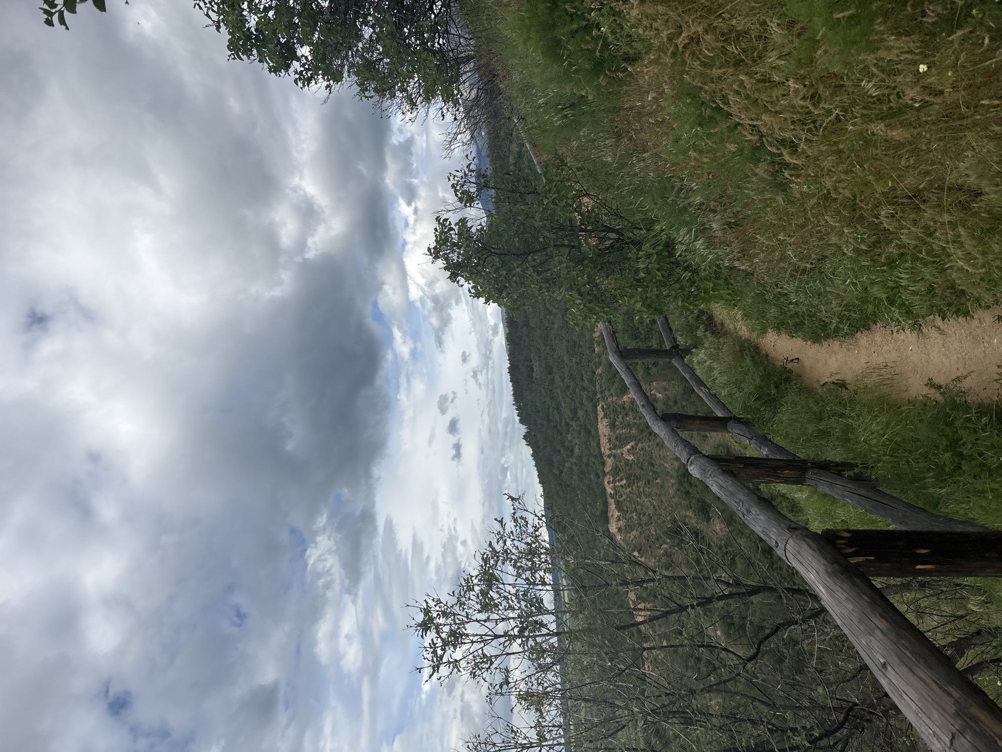 A medium-shot, low-angle landscape photograph depicts a dirt path bordered by a rough wooden fence, with a background of a cloudy sky and a hillside covered in vegetation. The path is composed of light-colored dirt and is located on the right side of the image, running diagonally away from the viewer. Adjacent to the path is a fence constructed from dark, roughly-hewn wooden posts and rails. The hillside, visible in the mid-ground, is covered in green vegetation, likely trees and shrubs. The sky occupies a significant portion of the frame and features a mix of grey and white clouds. Some trees are positioned in the foreground, near the bottom and top of the image, adding depth. The scene appears to be from a rural or natural setting. The location is Stob, Bulgaria.