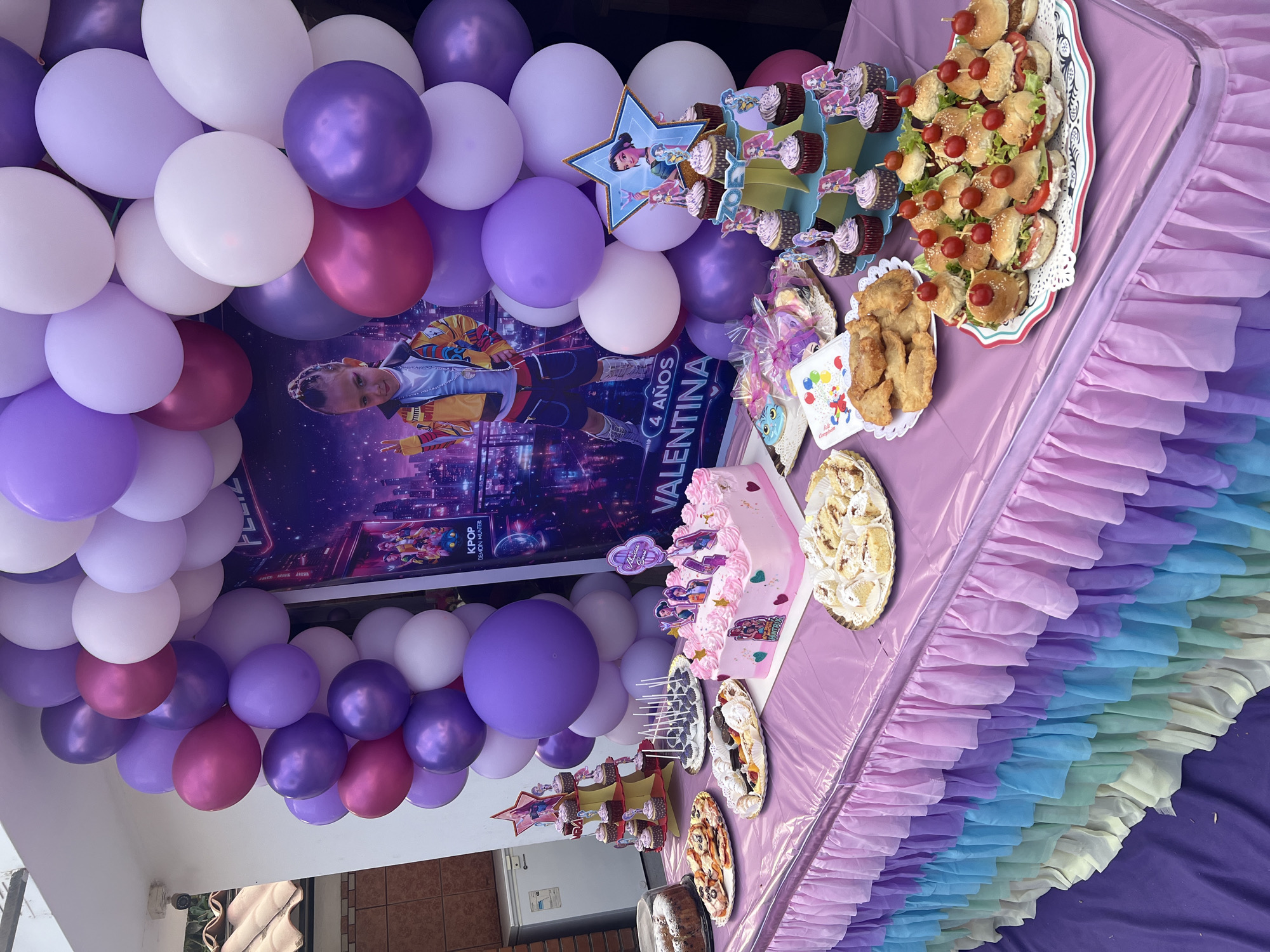 The image depicts a birthday celebration set up in Arica, Chile, likely for a child named Valentina who is turning 4. The scene is dominated by a decorated table laden with food. The table is covered with a light purple plastic tablecloth and edged with layers of colorful ruffled fabric.
Visible food includes sandwiches with tomatoes on top, a pink frosted cake adorned with candles and character cutouts, cupcakes on tiered stands, fried foods, various cakes and desserts displayed on platters, and what appear to be personalized treat bags.
A large poster with an image of Valentina and the words "4 años VALENTINA" is visible in the background. A balloon arch featuring white, light purple, dark purple, and pink balloons frames the poster. The event appears to be held outdoors, adjacent to a building with a white facade and a visible window. The backdrop suggests an urban setting, consistent with Arica. No individuals are clearly visible in the frame.
