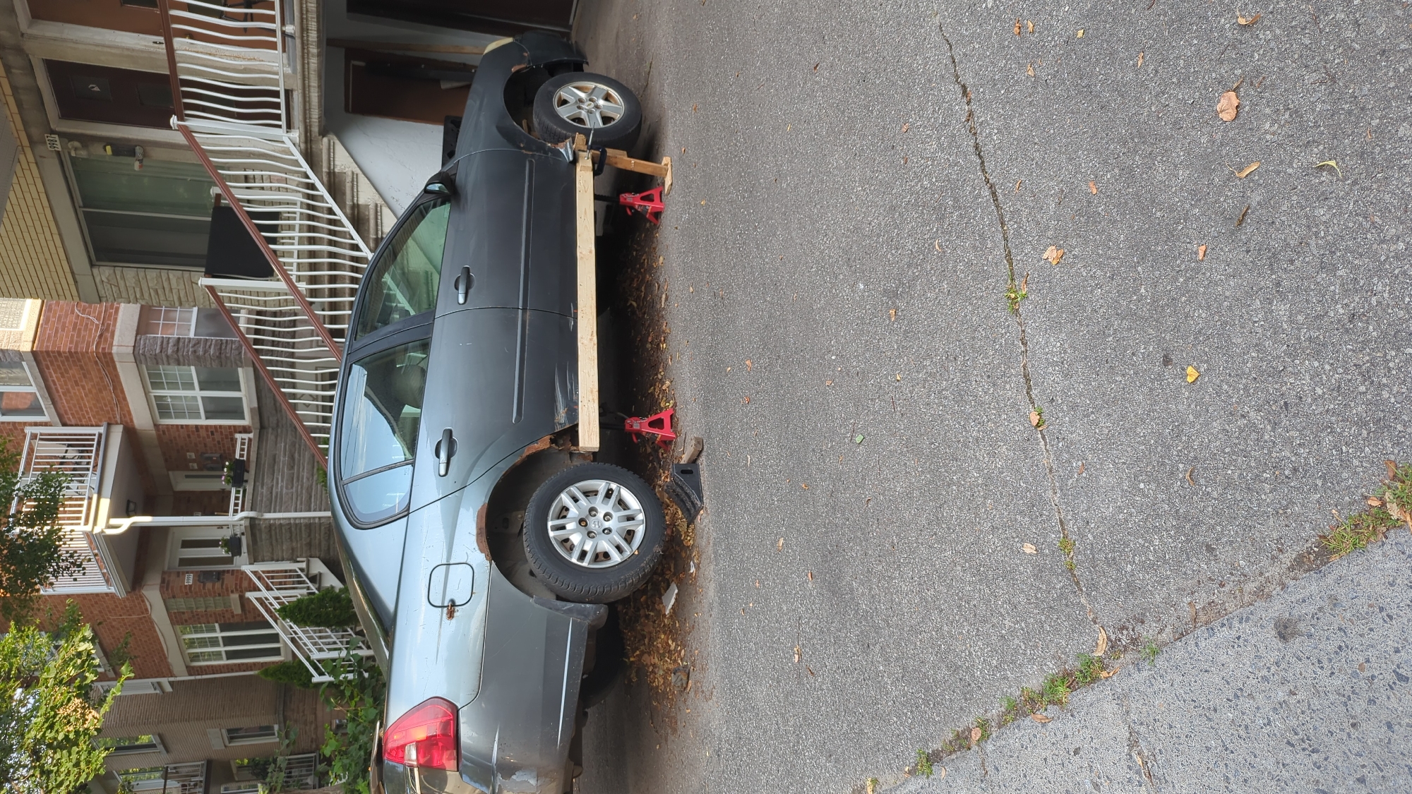 A damaged grey sedan is parked on a paved surface in front of residential buildings in Montreal, Canada. The car is elevated on four red jack stands and a wooden plank, indicating it is undergoing maintenance or repair. Rust is visible on the rear fender of the car. The residential buildings appear to be townhouses with brick facades and white balconies. The ground is covered with some fallen leaves, suggesting it might be autumn. The scene is outdoors in daylight.