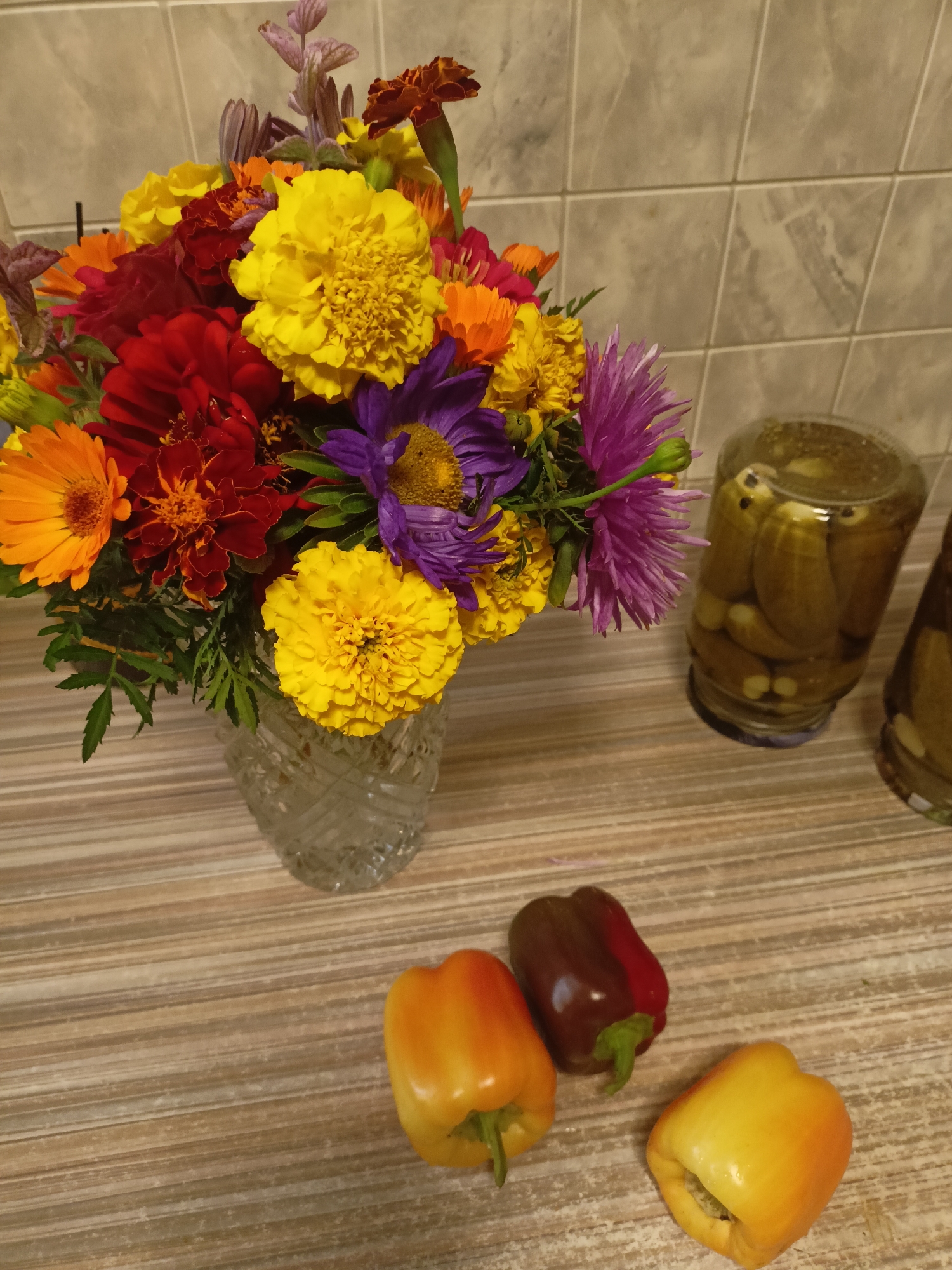 In an indoor setting, likely a kitchen, a crystal vase overflows with a vibrant bouquet of marigolds, asters, and other colorful flowers. The flowers are a mix of yellow, orange, red, and purple hues, with some buds and greenery adding depth. To the right of the flowers, two glass jars filled with pickled cucumbers stand on a tiled countertop. In the foreground, three bell peppers – two yellow and orange, and one dark red and red – are arranged on the same countertop. The countertop has a striped wooden pattern, and the wall behind is covered in light-colored tiles. The scene appears to be staged for a still life, with no people or animals present. The lighting is somewhat dim, suggesting it might be evening or a dimly lit room. The location is specified as Belorechensky, Russia.