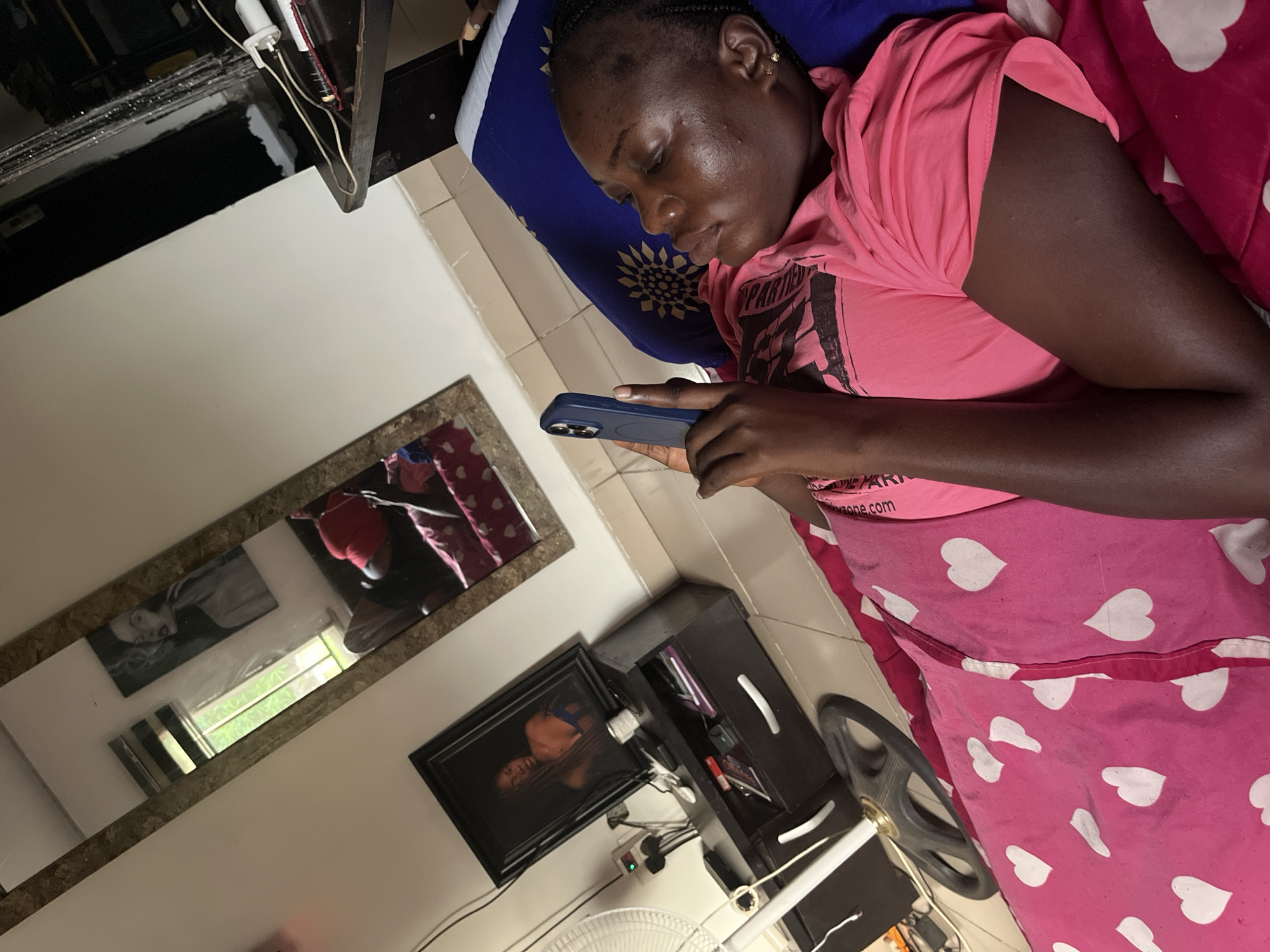 A dark-skinned individual, likely a young adult female, is depicted lying down indoors, fully absorbed in a dark blue smartphone held in both hands. Her hair is styled in braids, and she wears a small earring in her right ear. She is dressed in a pink t-shirt with partially visible text on it and is covered by a pink blanket or sheet adorned with white heart patterns, which also has "zone.com" printed along an edge. Her head rests on a blue pillow featuring a circular, sun-like design.
The setting appears to be a bedroom or personal living space, with light-colored tiled flooring. To the left, a long, ornate mirror hangs on a white wall, reflecting a bright outdoor scene with green foliage, indicating it is daytime. Within the mirror's reflection, another framed picture is visible on the wall. Below the mirror, a framed portrait of a person is placed on a dark wooden cabinet or dresser, which also holds various items and shows a white power adapter with wires. Part of a standing fan is visible in the bottom right corner of the frame. The overall scene suggests a moment of casual relaxation or focused engagement with a device.