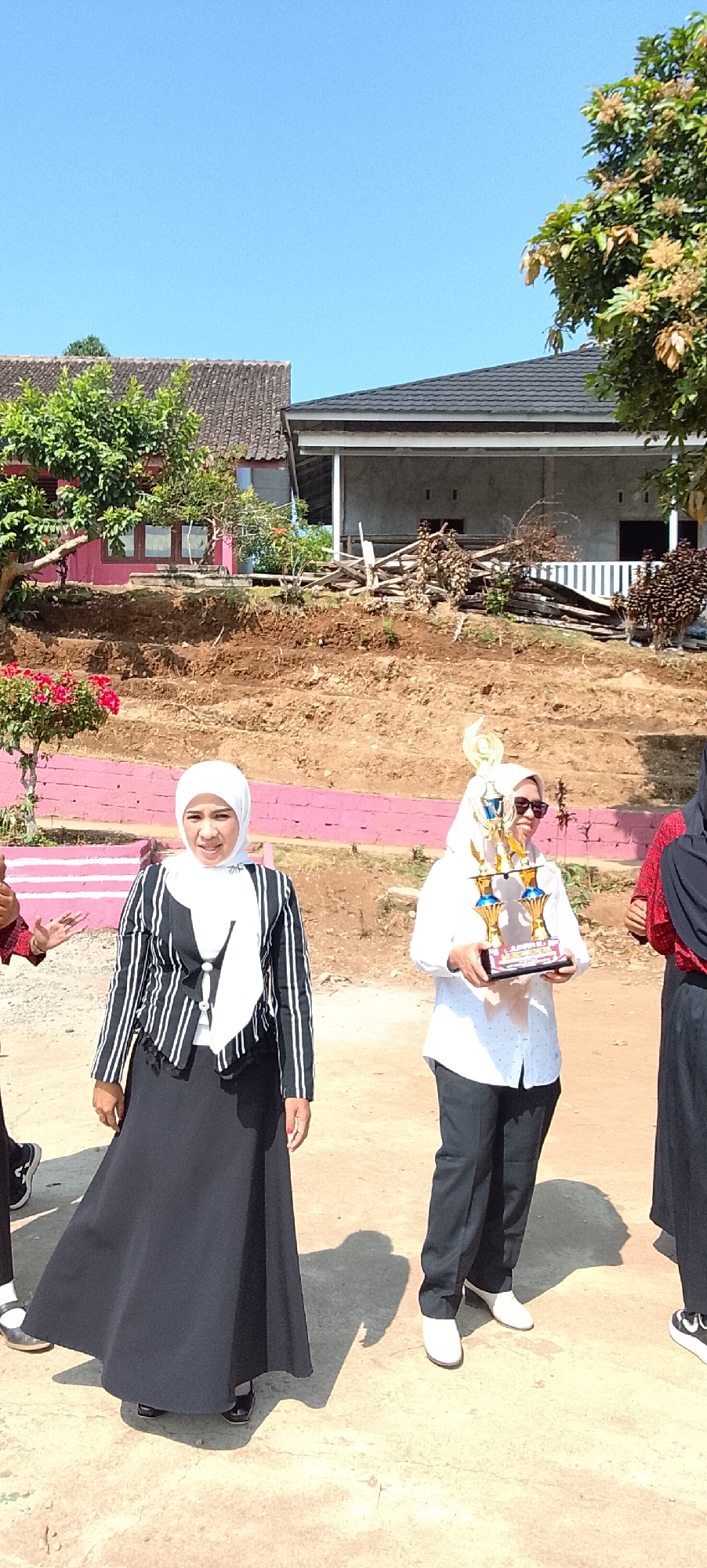 A group of women are standing outdoors in front of a house and a hill. The woman on the left is wearing a black and white striped jacket and a long black skirt. She is smiling and looking at the camera. The woman in the center is wearing a white shirt, black pants, and a white hijab. She is holding a trophy and smiling. The woman on the right is wearing a black hijab and a red and black patterned shirt. She is partially visible. In the background, there is a house with a pink facade and a grey roof. A hill behind the house is covered in dirt and some debris. The sky is clear and blue. The image appears to have been taken during the day. The location is not Jepara, Indonesia.
