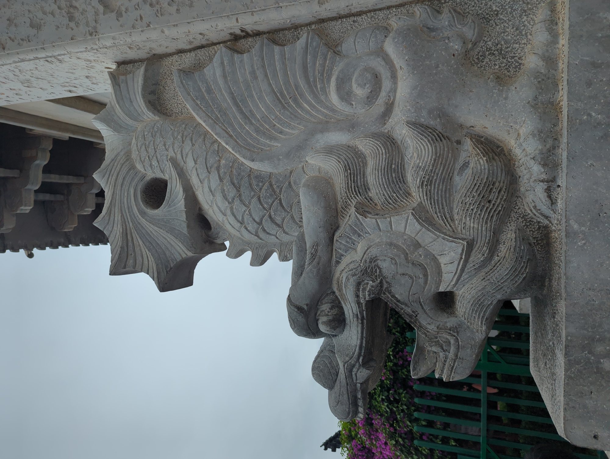 This media file features a close-up, low-angle shot of intricately carved stone ornamentation, likely part of a traditional Chinese architectural element. The carving depicts a mythical creature, resembling a dragon or a Qilin, with scales, elaborate fins, and a fierce, stylized head. The detail in the carving is evident, showcasing textured scales, flowing mane-like features, and sharp teeth. The stone itself appears weathered, suggesting it is an older structure.
The background is dominated by a hazy, overcast sky, indicating a cloudy or foggy day. Below the stone carving, a glimpse of lush green foliage with vibrant purple flowers can be seen, suggesting an outdoor setting with vegetation. A dark, slatted fence is also partially visible in the background. The perspective is from below, looking upwards at the architectural detail, which is mounted on a larger stone structure. The overall lighting is soft due to the overcast sky. There is no visible text in the media file. The scene evokes a sense of historical artistry and cultural significance.