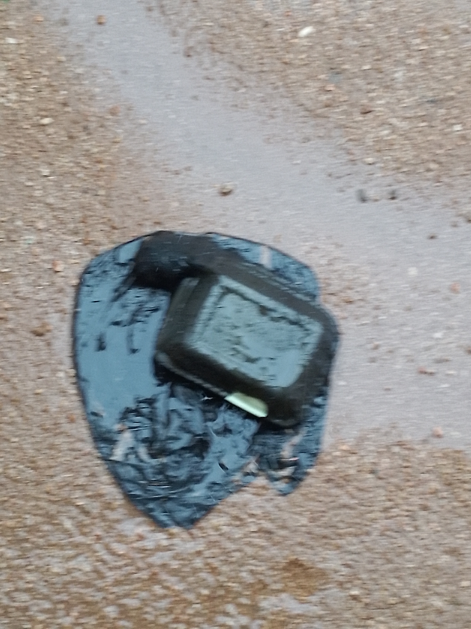 A black remote control is lying in a puddle on a gravelly surface. The remote is partially submerged in dark, wet material, possibly plastic sheeting. The surface appears to be an outdoor area, with small pebbles and dirt visible. The lighting is dim, and the image is slightly out of focus.