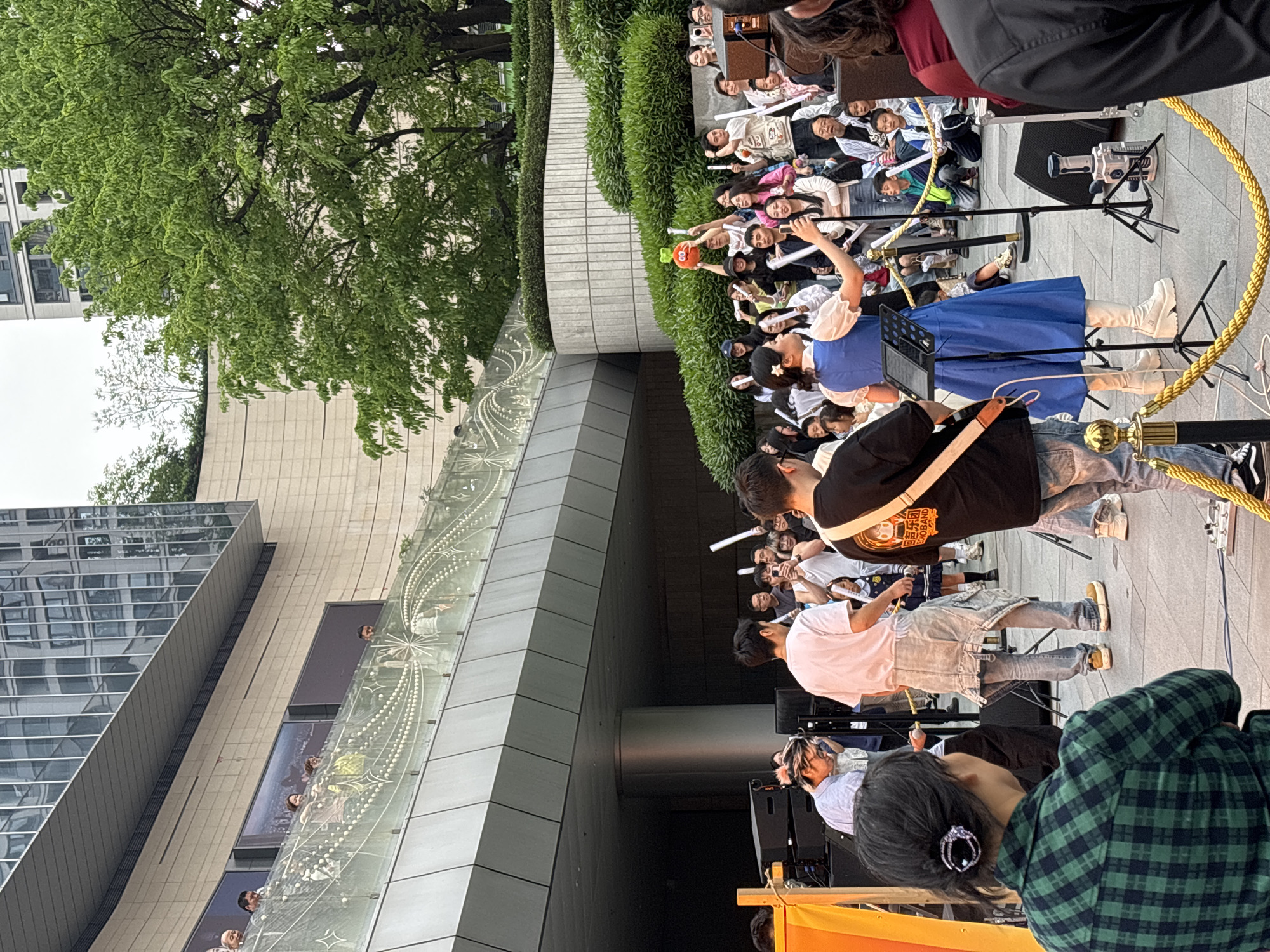 The image shows a live performance taking place outdoors in Jiulongpo District, China, with a crowd of spectators. A band, consisting of at least three members, is performing on a stage. The singer, wearing a blue dress and white boots, stands in front of a music stand. The spectators are holding what appear to be light sticks. A yellow rope demarcates the stage area. The backdrop features a modern building with reflective glass and design elements. A large tree is situated to the side of the building. The scene is set during the daytime, presumably in an urban environment. Some people are watching from higher vantage points within the building.