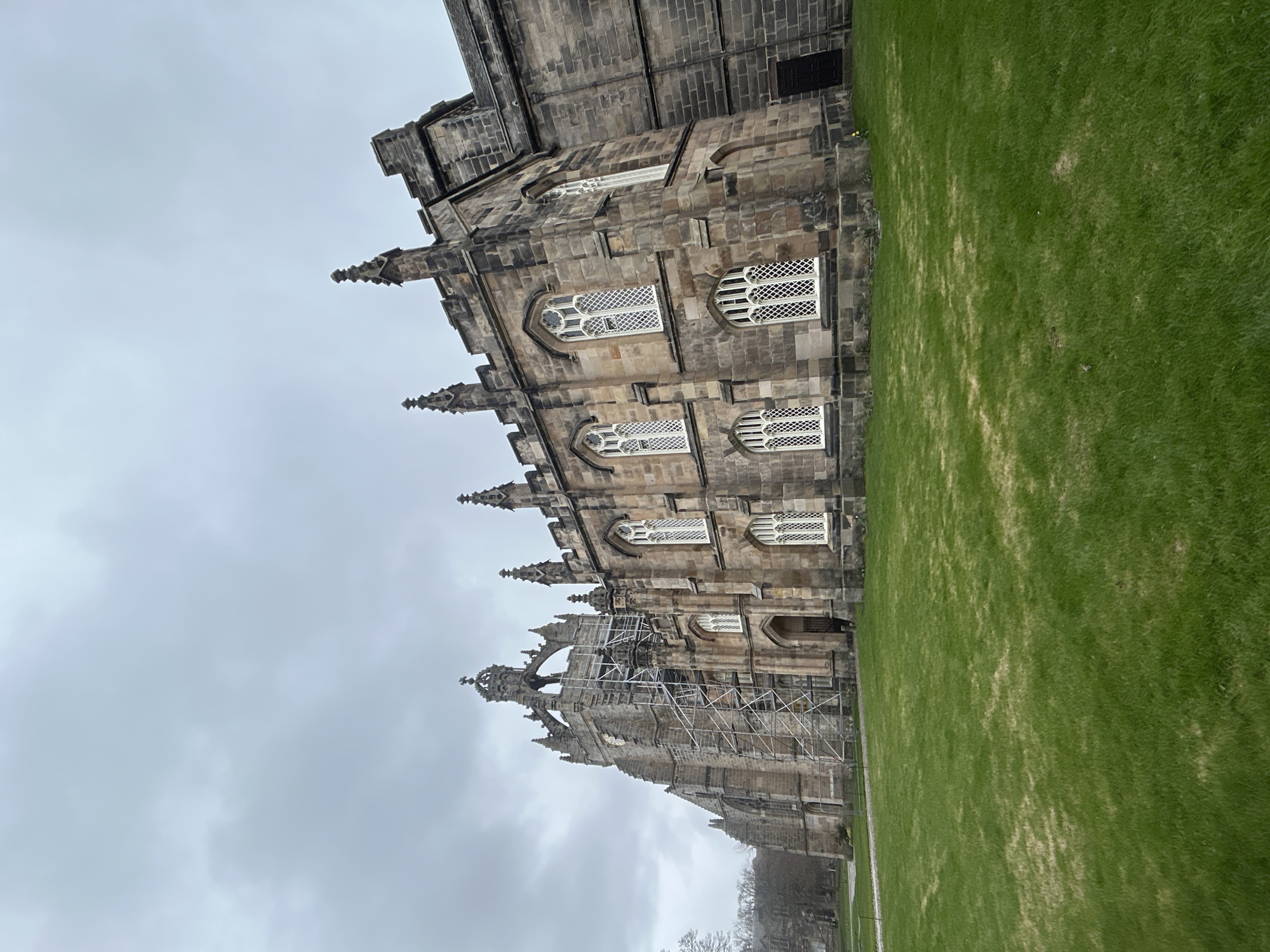 The image shows a large, ornate stone building with Gothic architectural elements, including spires, pointed arches, and decorative windows. The building is likely a historic structure, possibly a university or church. Scaffolding is erected on one section of the building, indicating ongoing maintenance or restoration work. The building is adjacent to a grassy lawn. The sky is overcast, suggesting a cloudy day. The location is Aberdeen City, United Kingdom.