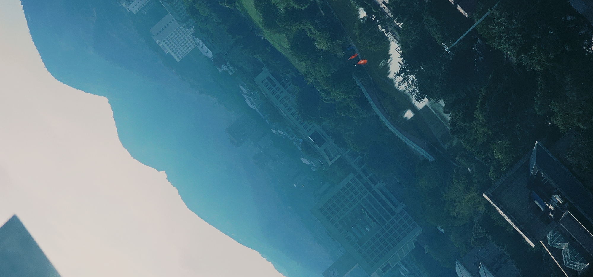 The image, taken at an oblique angle, depicts an aerial view of a cityscape with a prominent mountain range in the background. Buildings with distinct architectural features, including grid-like windows and multiple stories, are interspersed with dense green foliage. A body of water, likely a lake or river, is visible with a bridge arching over it. Two red flags are noticeable near the water's edge. The overall scene suggests an urban environment with significant natural elements, possibly a park or a university campus, located in Chongqing High-tech Zone, China. The lighting suggests it is daytime, with a hazy or overcast sky.