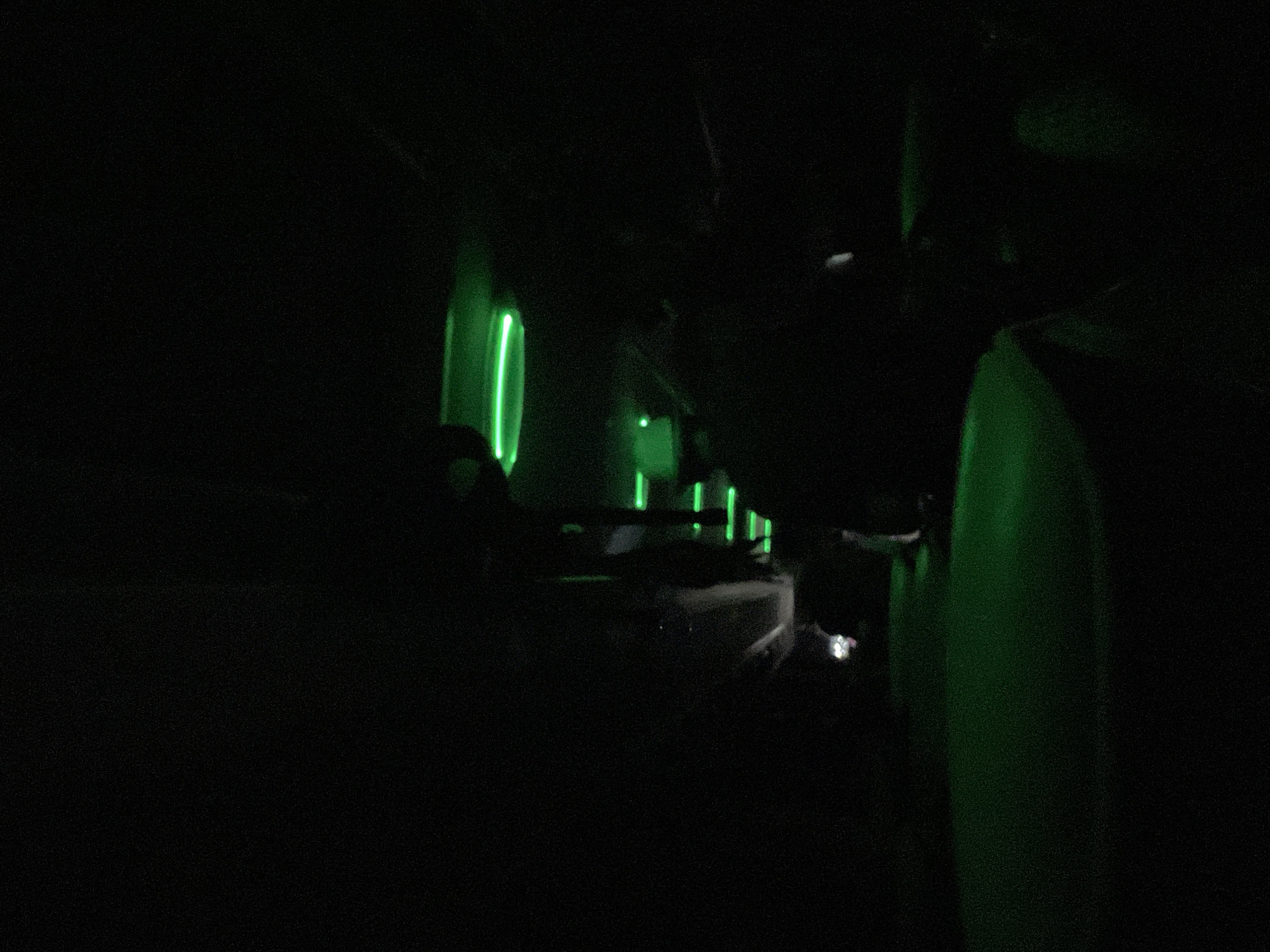 The image appears to be taken inside an airplane cabin at night. The scene is dimly lit with green accent lights. In the foreground, part of an airplane seat is visible. Further back, a row of windows illuminates green light inside of the cabin. There are people sitting in their seats. The location is unknown. There are no indications of Kalugerovo, Bulgaria.