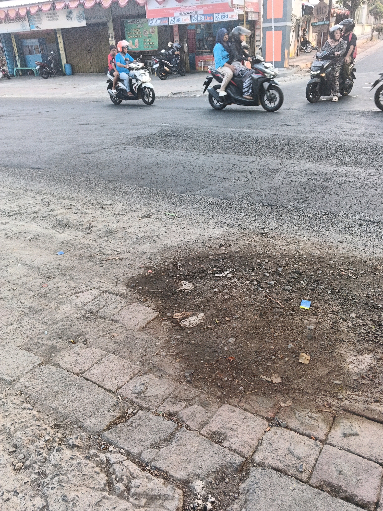 The image captures a street scene in Ungaran, Indonesia, depicting several motorbikes and their riders. In the foreground, a white motorbike carries two individuals, one wearing a blue shirt and orange helmet, and the other a red shirt. They are traveling on a paved road with visible cracks and debris. To the left of this motorbike, another motorbike is parked. Further down the street, a dark-colored motorbike with two passengers, both wearing helmets, is in motion. Adjacent to it, another dark motorbike also has two occupants. The background shows buildings, suggesting a commercial or residential area. The street appears to be a typical urban environment with varying road conditions.