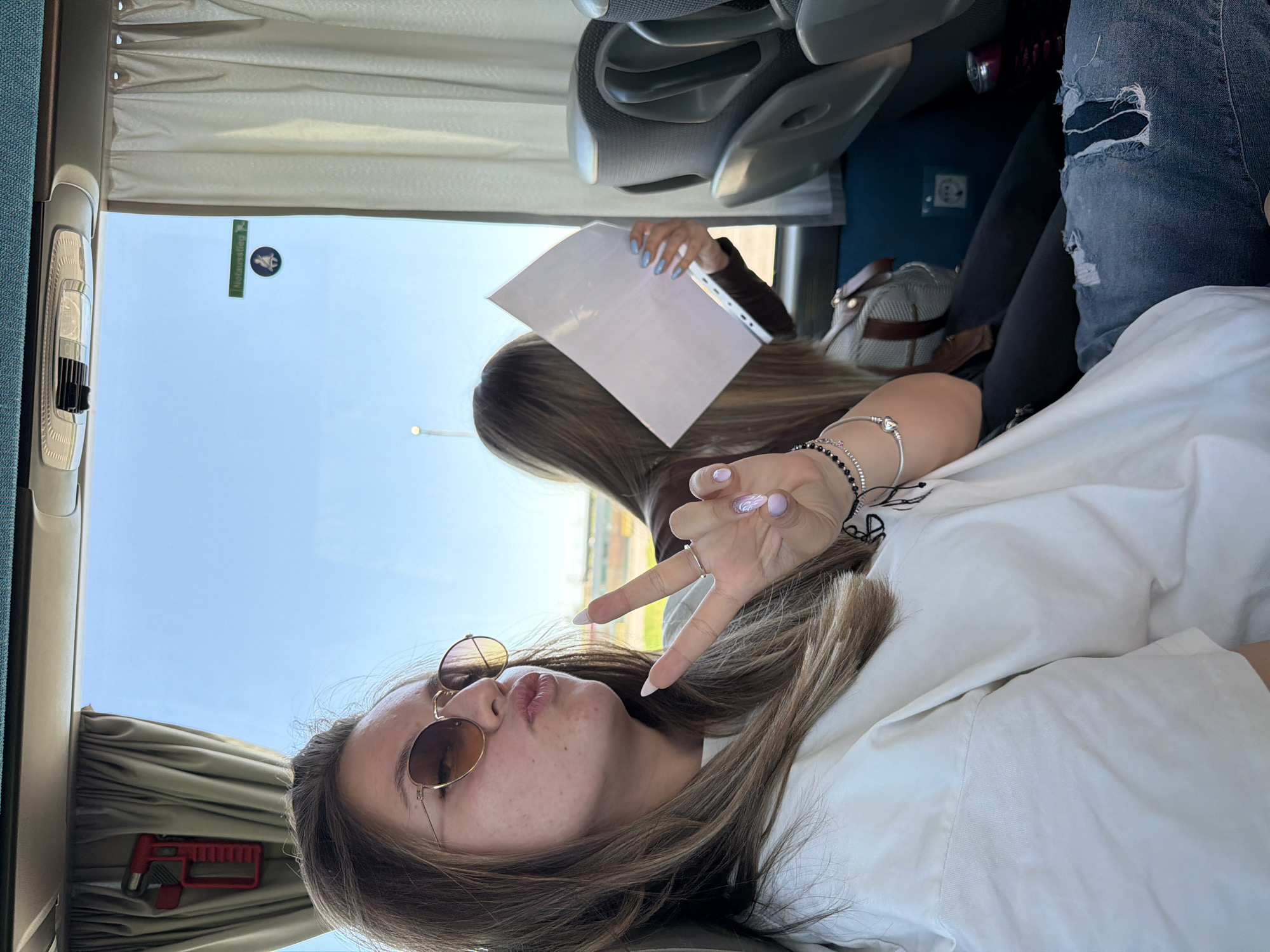 The image shows a woman with brown hair and sunglasses making a peace sign in what appears to be a bus. She is wearing a white shirt. Another person is visible in the background holding a white sheet of paper. The bus window shows an exterior view; an airport is visible. The bus is in Kapitan Andreevo, Bulgaria.