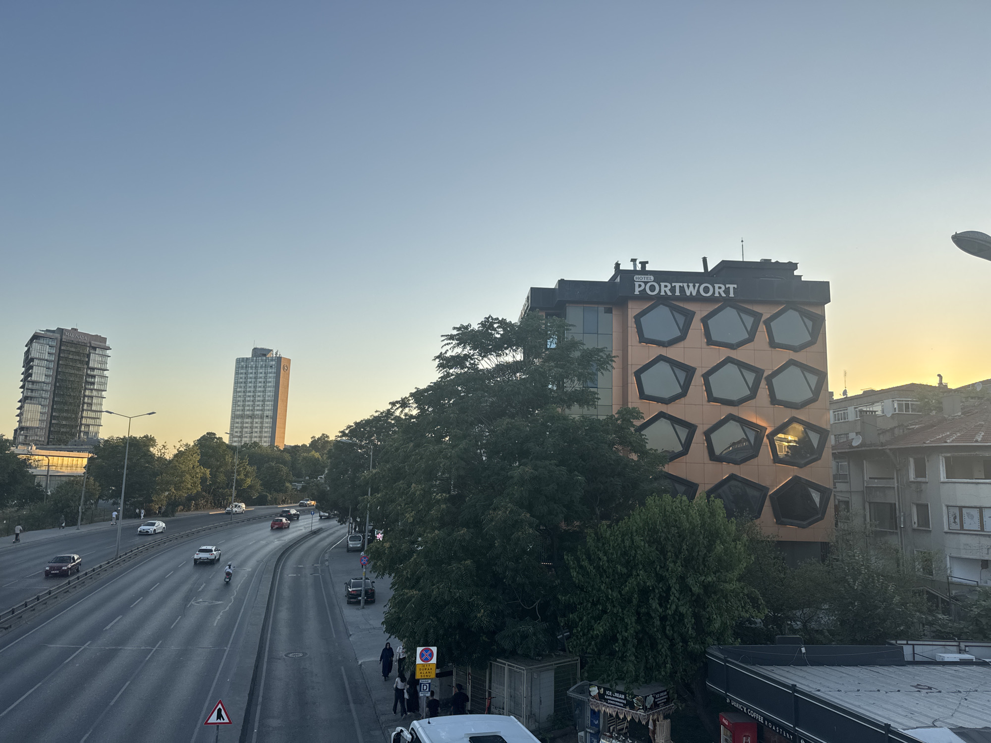 The image captures a street scene in Bakırköy, Turkey, during what appears to be late afternoon or early evening, with the sun low on the horizon. In the foreground, a multi-lane road with moderate traffic is visible, including cars, a scooter, and a motorcycle. Pedestrians are seen on the sidewalks.
Dominating the mid-ground is a modern building with a distinctive facade featuring an arrangement of irregularly shaped, dark-framed windows on an orange-colored surface. The building is labeled "HOTEL PORTWORT". Adjacent to it, a dense cluster of trees provides a natural element.
In the background, two tall, modern buildings with reflective glass exteriors rise against the clear, pale blue sky. Further to the right, older, lower-rise residential buildings are visible, some showing signs of wear. A street lamp with a curved arm is present on the right side of the image. The overall impression is of an urban environment with a mix of architectural styles, ongoing traffic, and people going about their day.