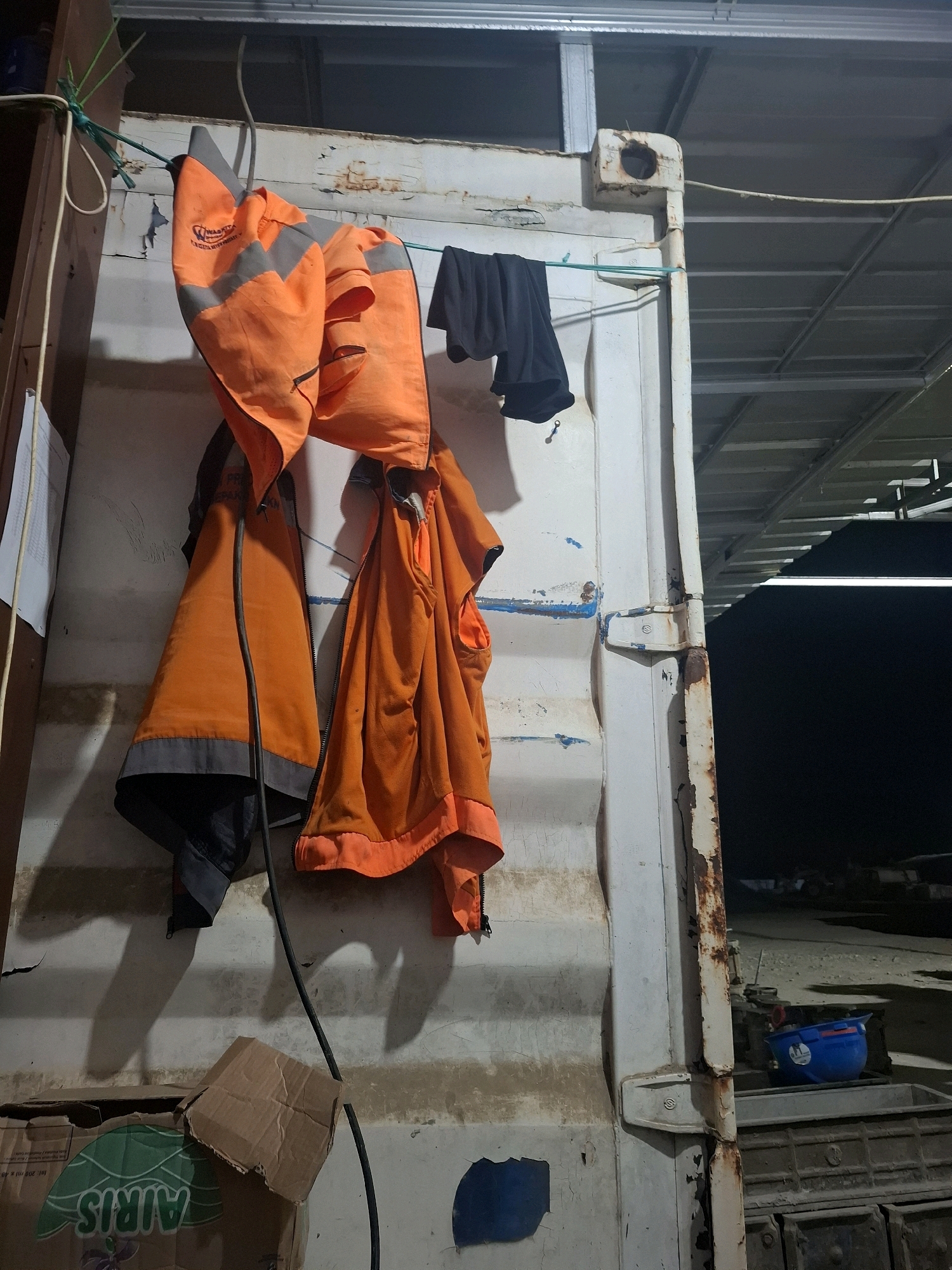Two orange safety vests and a pair of black shorts are hanging on a clothesline strung across the side of a white shipping container. The vests have reflective stripes. To the left of the container, a piece of white paper is attached to a wooden support. Below the vests, a dark cable hangs down. On the ground in front of the container, there is a flattened cardboard box and another cardboard box with the word "AIRIS" visible on it. In the background, a blue bucket and a grey bin are visible. The scene appears to be outdoors, with a covered structure in the background. The location is specified as Kutasirna, Indonesia.