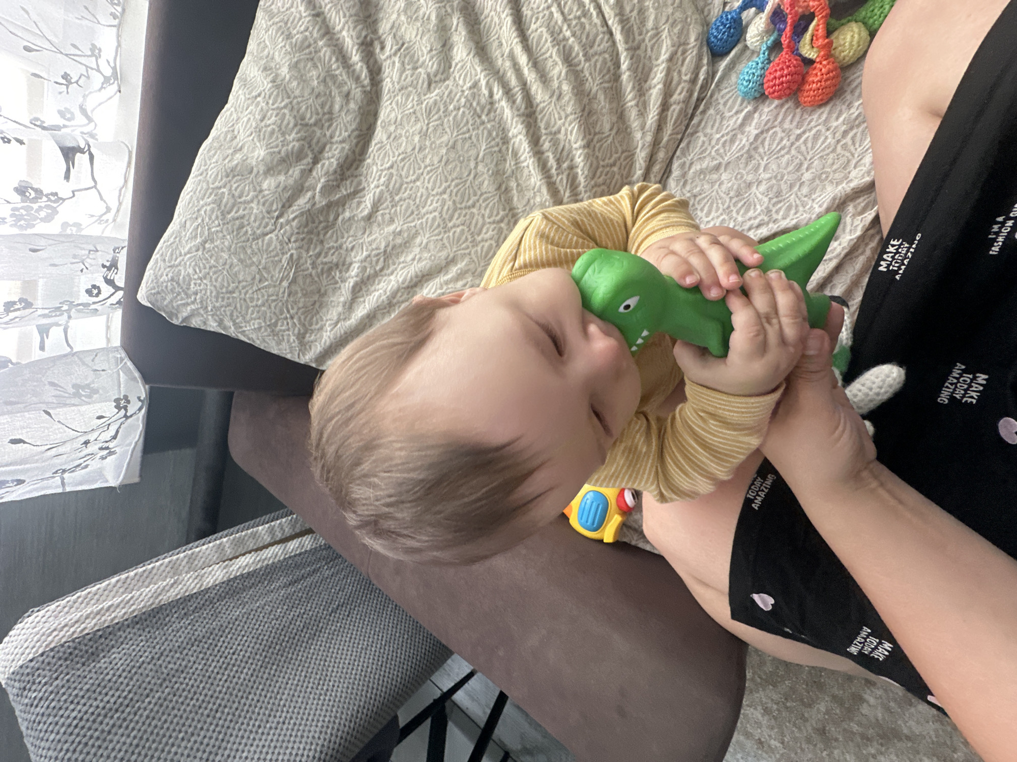 A close-up shot shows a baby boy with light brown hair lying on a patterned blanket. He is wearing a yellow and white striped long-sleeved shirt and is holding a green toy dinosaur up to his face. The baby appears to be sleeping or very relaxed, with his eyes closed. Next to him, a person's arm and leg are visible, wearing black shorts with white text that reads "MAKE TODAY AMAZING." In the background, a white curtain with a branch pattern is visible, as well as a stack of colorful crocheted balls. The scene appears to be indoors, likely in a home, with soft lighting. The overall mood is peaceful and comforting.