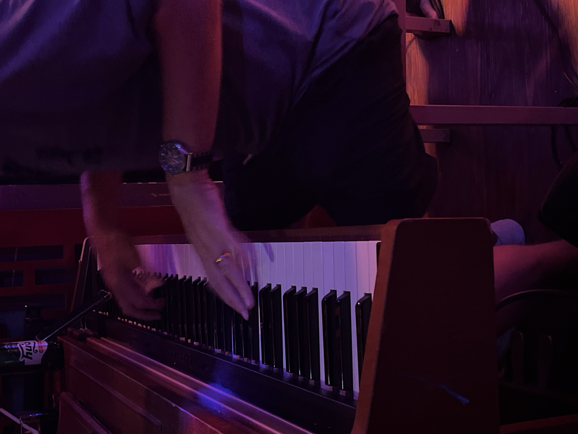 A person is playing a piano. The person is wearing a watch on their left wrist and a ring on their left hand. The piano is brown and white. There is a can of beer next to the piano. The location is likely a bar or performance venue. Đà Nẵng, Vietnam is not discernible from the image.