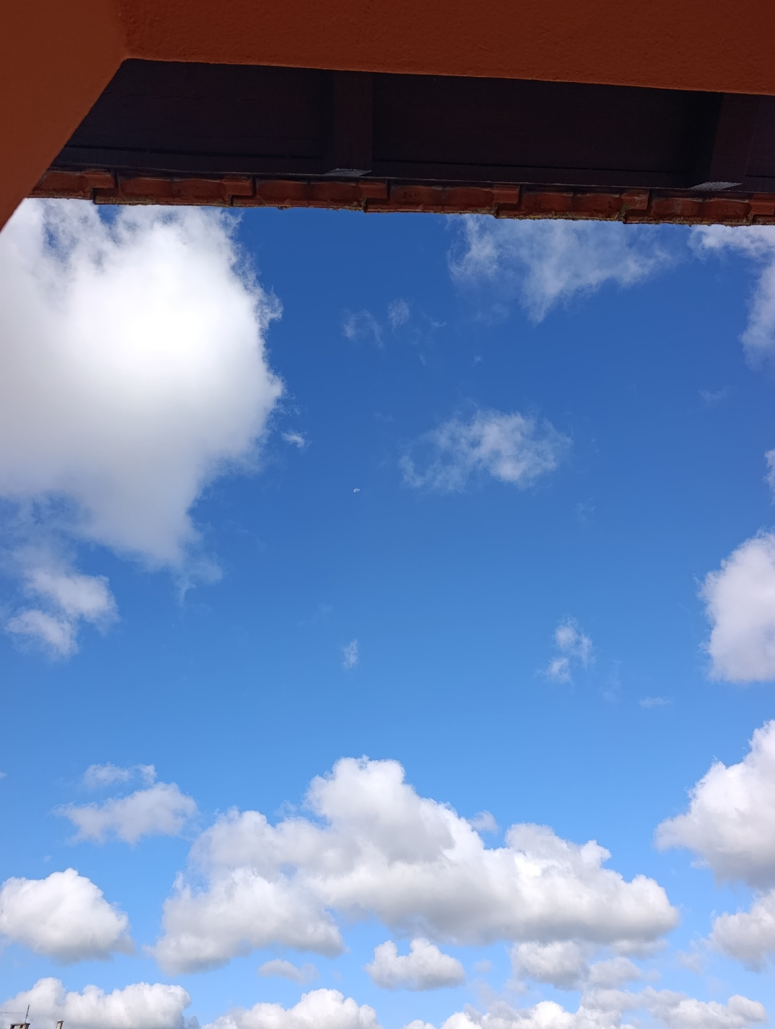 A low-angle shot looks up at a bright blue sky filled with scattered white clouds. A small, white object, possibly a distant airplane or drone, is visible in the upper right quadrant of the sky. The top of the frame is partially obscured by a reddish-orange overhang and dark wooden beams, suggesting a rooftop or balcony structure. The edge of a brick wall is visible just below the overhang. The scene appears to be an outdoor urban environment, likely Setúbal, Portugal, given the architectural style and sunny sky. There are no people or animals visible in the frame, and no discernible actions or interactions are taking place. The overall impression is one of a clear day with a vast sky.