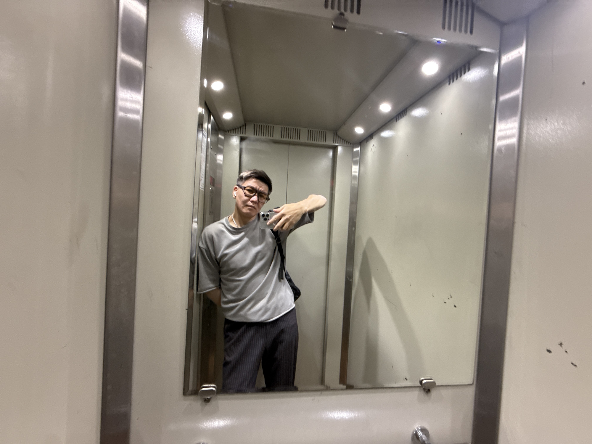 A man takes a mirror selfie inside an elevator, reflecting his image in a large rectangular mirror. He is positioned slightly to the left, wearing a grey short-sleeved t-shirt, dark pinstriped trousers, and rectangular glasses. A black bag strap is visible over his right shoulder, and he has a white earbud in his left ear. He holds a smartphone with his left hand, angling it towards the mirror to capture the photograph. His expression is neutral to slightly serious.
The elevator interior features light-colored, possibly off-white or cream, walls and ceiling. Multiple recessed spotlights illuminate the space brightly. Metallic, likely stainless steel or chrome, vertical panels frame the mirror and are also visible as part of the elevator doors reflected in the background. Ventilation grates are noticeable on the ceiling and along the upper part of the reflected walls. The mirror itself shows some smudges and marks, and the surrounding wall has a few small dark spots. The overall setting is an indoor elevator, consistent with a building environment in Moscow, Russia. There is no visible text in the image.