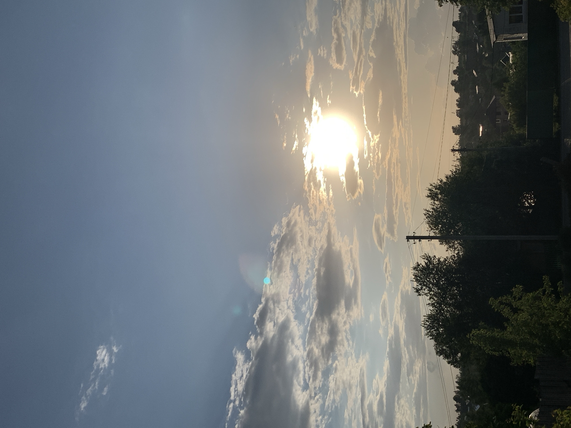 The image shows a sky scene with the sun obscured by clouds. The left side of the image features a clear blue sky with some scattered clouds, while the right side depicts dense clouds around the sun, creating a bright, diffused light. The horizon line reveals a silhouette of trees, buildings, and utility poles, suggesting a residential area. Based on the utility poles and building details, this area could be identified as the city of Фоки, Russia.