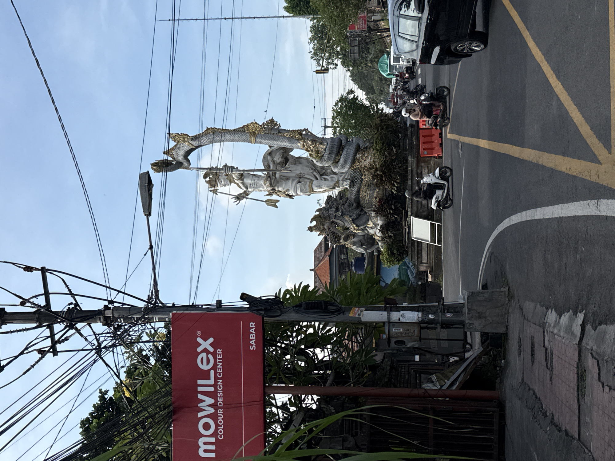 This media file captures a vibrant street scene, likely at an intersection, in Ubud District, Indonesia, during bright daylight hours. The sky is clear blue, indicating sunny weather.
At the center of the image stands a large, ornate stone statue, predominantly white or light grey with gold accents. It depicts a deity or mythical figure, possibly holding a bow and arrow, intricately carved with flowing garments and a headdress. A prominent, large serpent or dragon-like creature is entwined around the statue, also detailed with gold. This statue appears to be situated on a small median or island in the road.
The surrounding area is active with daily traffic. On the asphalt road, painted with distinctive yellow and white markings (including a large V-shaped yellow line), several motorcycles and scooters are visible, some with riders. A black car is also parked or moving on the right side of the road. Red concrete barriers are seen near the roadside, possibly for traffic management or construction.
Overhead, a dense and complex network of black power lines and cables crisscrosses the sky, attached to utility poles. A street light fixture is also visible among these wires. To the left, a prominent red rectangular sign is attached to one of the utility poles. The text on this sign reads "MOWILEX" in large white letters, with "COLOUR DESIGN CENTER" underneath, and "SABAR" in a smaller red box below that.
Lush green trees and foliage are scattered throughout the background, contrasting with the urban infrastructure and hinting at the district's natural beauty. Buildings with traditional-style roofs can be glimpsed behind the statue and greenery. The overall impression is one of a busy, yet culturally rich, street environment typical of the region.