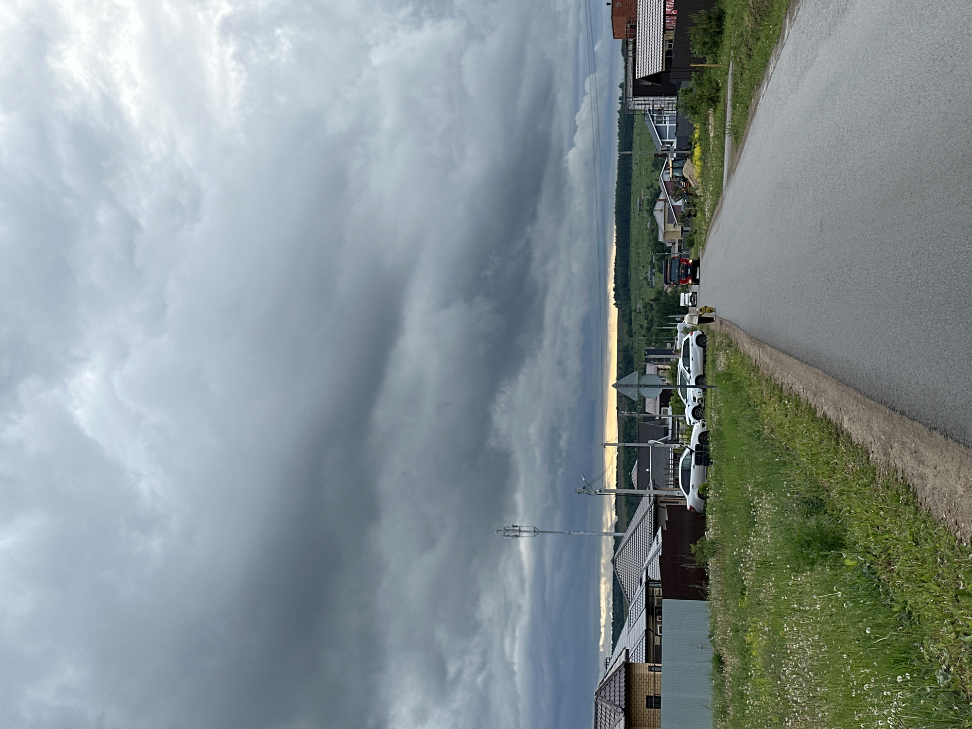 A roadside view in Эстачи, Russia, shows an asphalt road on the right, bordered by a grassy verge filled with dandelions. Several white cars are parked along the edge. A row of houses stands on the left, interspersed with telephone poles. In the distance, a glimpse of trees can be seen. Above, a sky filled with heavy, gray clouds dominates the scene.