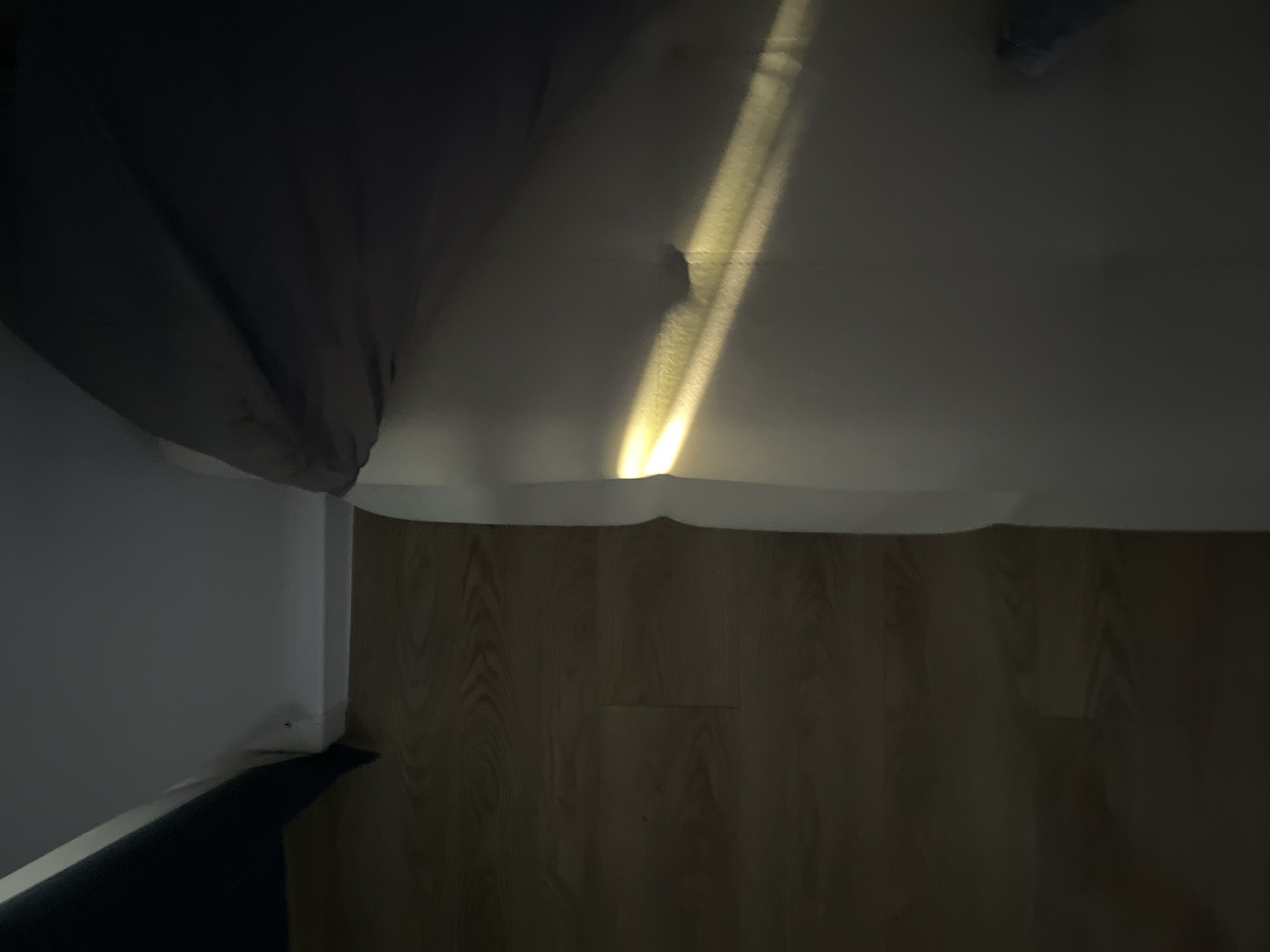 The image depicts an interior room with a laminate wood floor. A wall is visible on the left side of the frame, and a dark-colored drape or curtain hangs partially in the upper left corner. A beam of light shines across the ceiling. The location in Khimki, Russia is not determinable from the image.