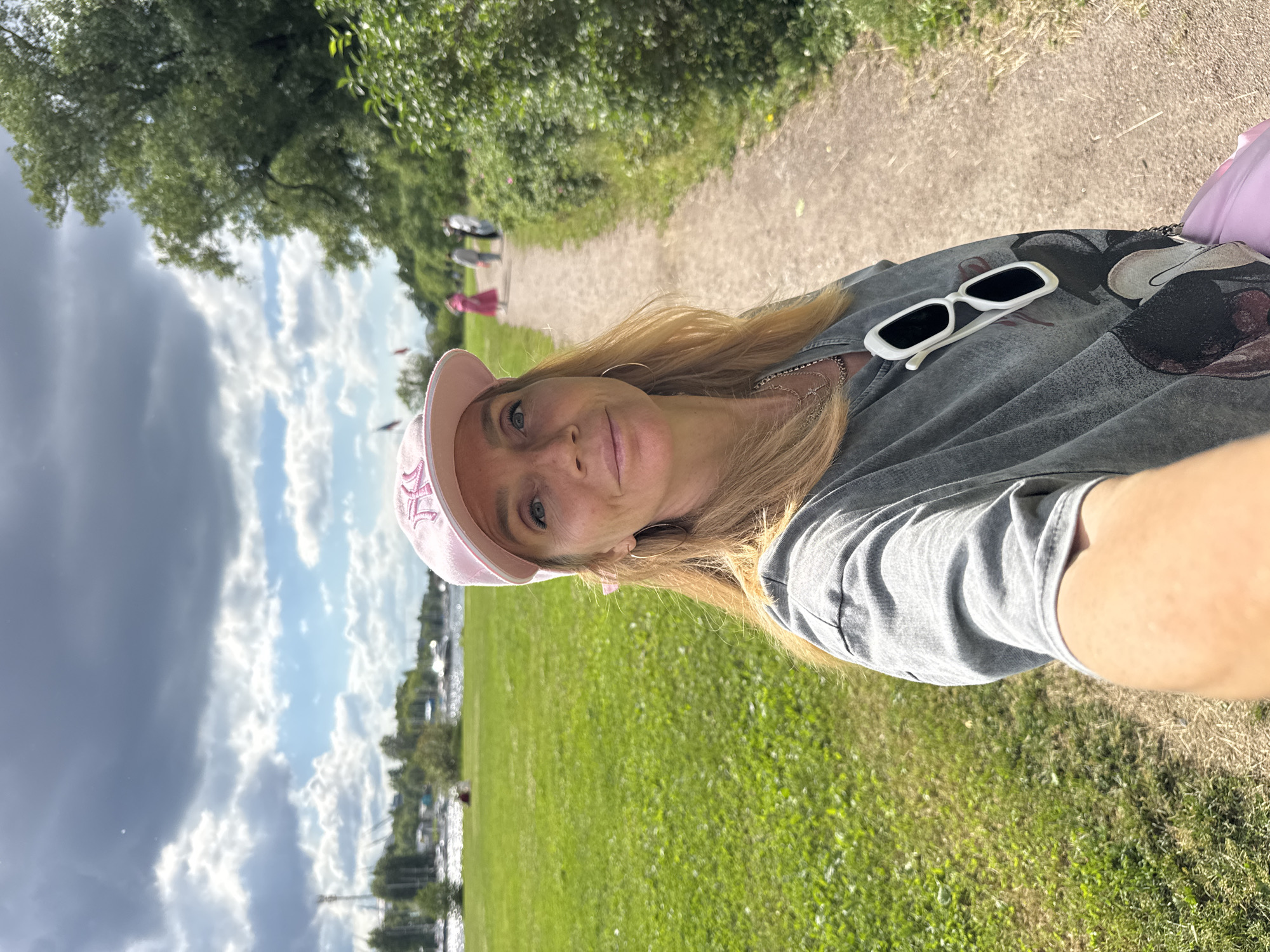 A woman with blonde hair and blue eyes takes a selfie while walking on a path in a park. She is wearing a gray t-shirt with a graphic print, a pink baseball cap, and sunglasses clipped to her shirt. She also has a silver necklace and hoop earrings. In the background, there is a grassy field, trees, and a body of water with boats. Several other people are walking on the path in the distance. The sky is cloudy with patches of blue. The location is identified as a park in Saint Petersburg, Russia.