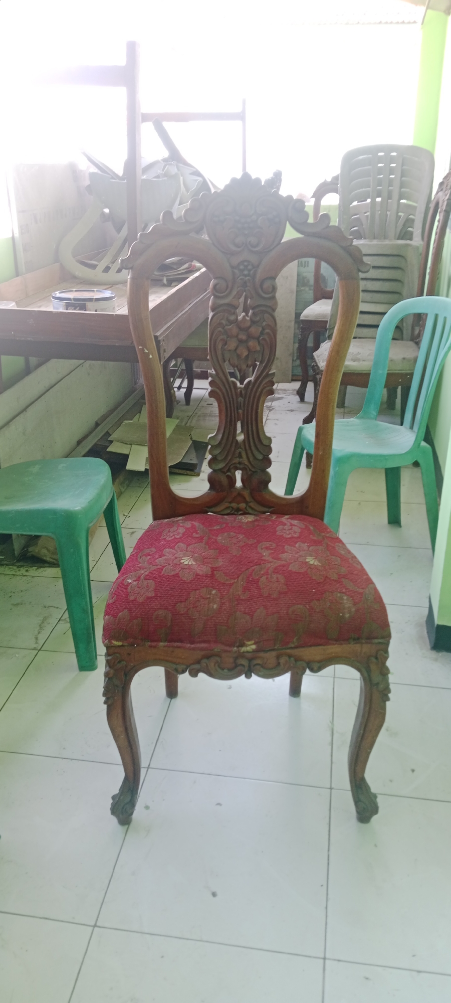 The image shows an ornate wooden chair with a red, patterned upholstered seat in the foreground. The chair has carved detailing on its back and legs. To the left of the chair is a green plastic stool. In the background, there is a wooden table with some items on it, and several plastic chairs are stacked or visible. The scene appears to be indoors, possibly in a workshop or storage area, with a tiled floor and bright light coming from a window or opening on the left. The overall impression is one of a cluttered or semi-finished space. The image location is identified as Tuban, Indonesia.