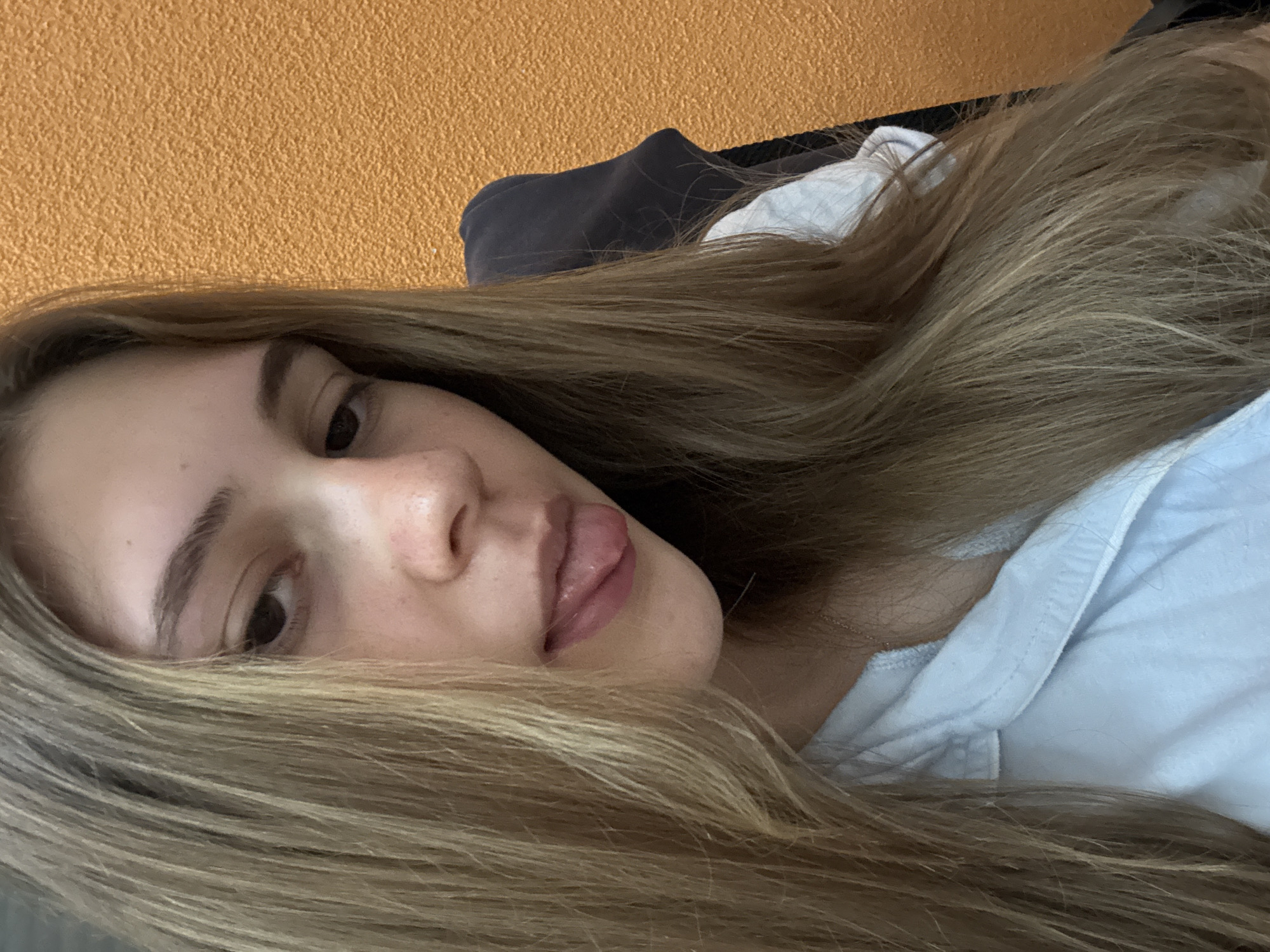A young woman with long, light brown hair is sticking her tongue out slightly. She is wearing a light blue shirt. The background is an orange wall. A dark grey object is also visible behind her. There is no information in the image to suggest this was taken in Panagyurishte, Bulgaria.