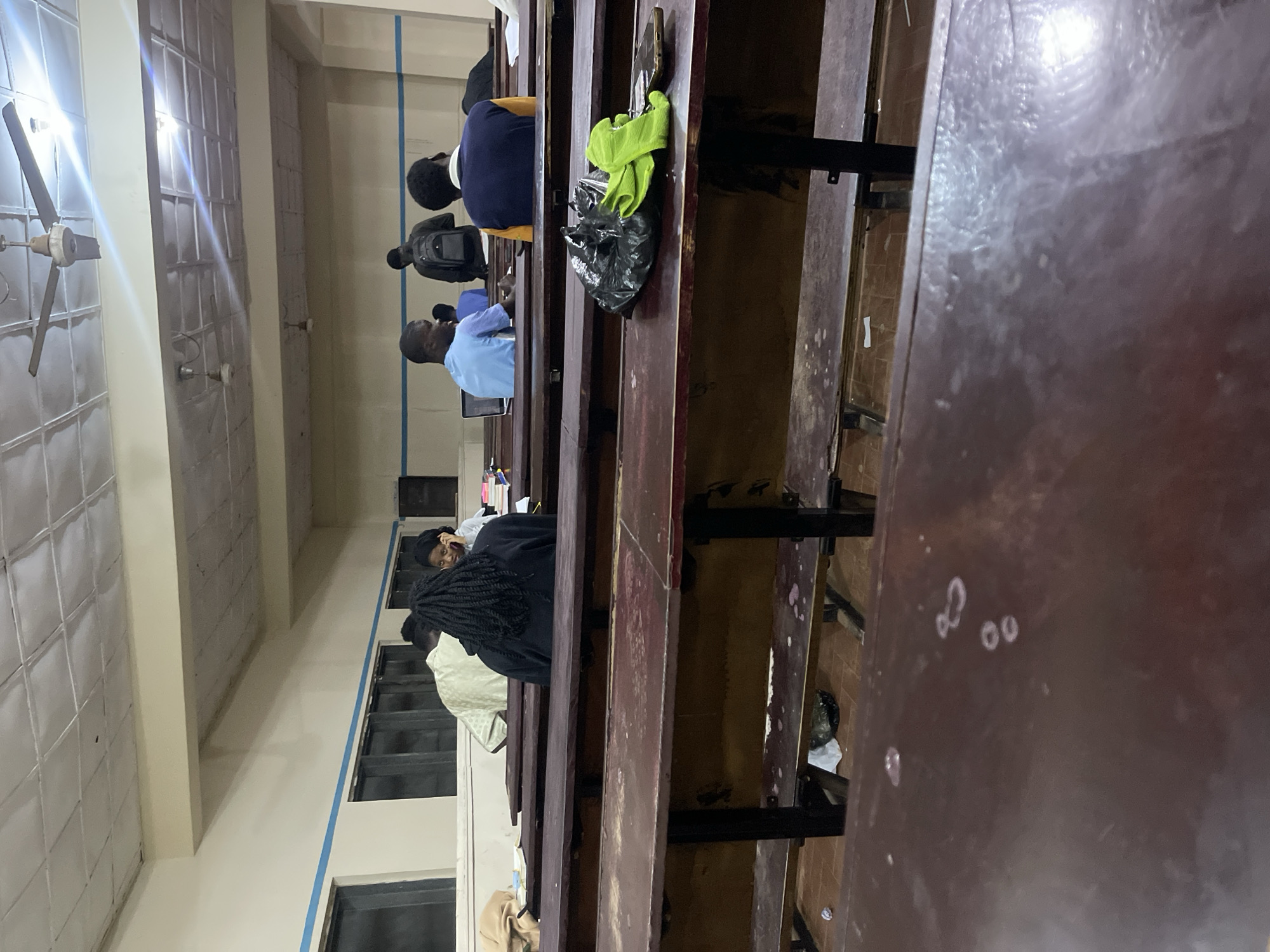 The image depicts an indoor scene, likely a classroom or lecture hall, with rows of wooden desks. In the foreground, a dark wooden desk surface is visible, showing some wear and tear. Further back, several individuals are seated at desks, facing away from the camera, suggesting they are engaged in a lecture or study session. One person in the middle ground is wearing a light-colored, patterned garment. To their left, another person with dark braided hair is seated, and further to the right, a person in a blue shirt is visible. In the background, there are more desks and individuals, as well as ceiling fans and fluorescent lighting. The overall environment appears to be a functional, utilitarian space. The provided location information, "Rafin Par, Nigeria," suggests this scene is situated within a community or educational institution in that region.