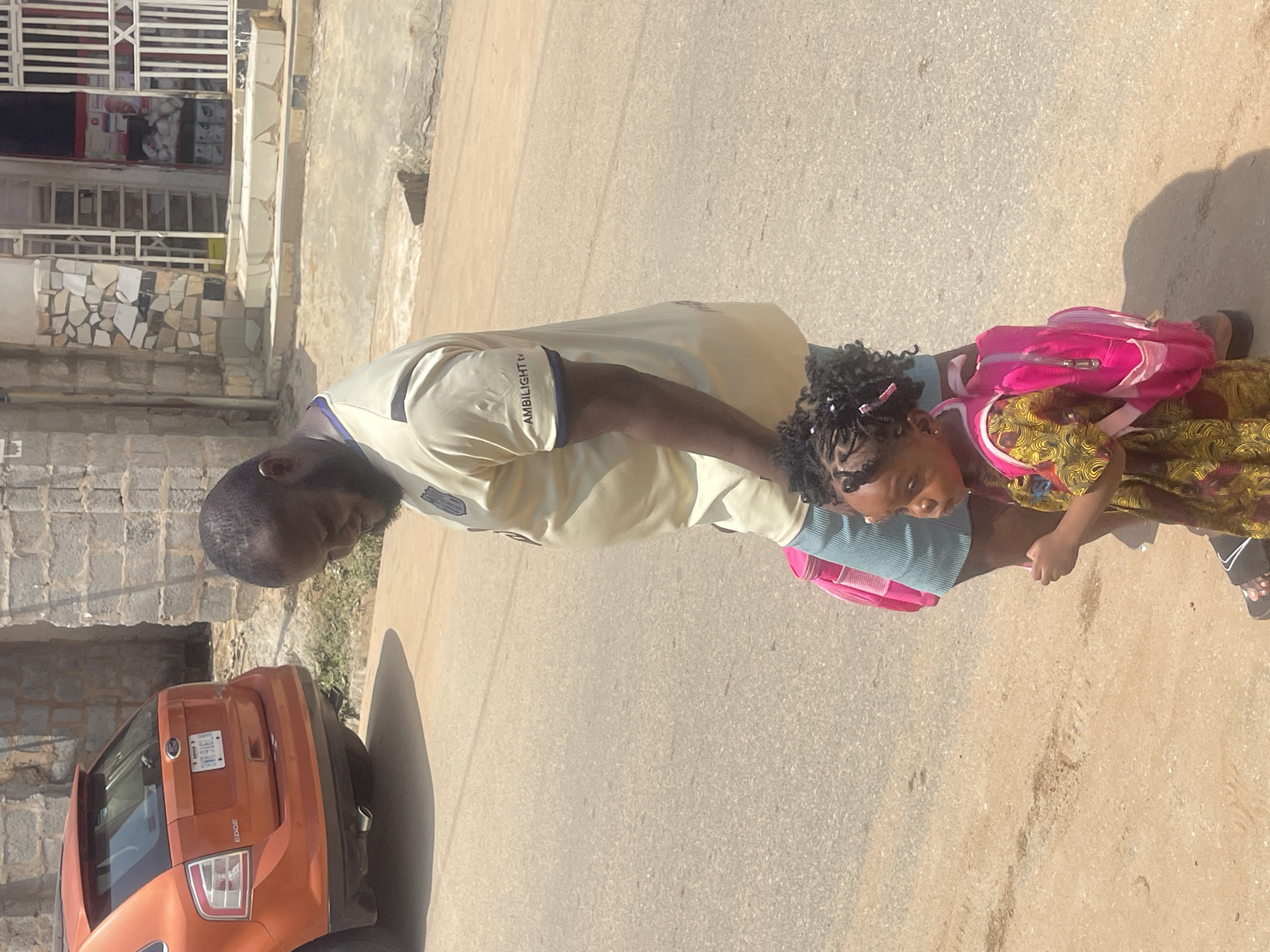 In Abuja, Nigeria, a man and a young girl are walking on a paved road. The man, wearing a light-colored shirt with "AMBILIGHT TV" printed on the sleeve, is positioned behind and to the left of the girl. The girl, dressed in a colorful patterned dress, is carrying a bright pink backpack. To the left of the pair, an orange SUV is parked. The background shows a building constructed with concrete blocks, stairs leading up to an entrance, and a street-level shop with a white metal gate. The overall scene depicts an outdoor urban environment under daylight.
