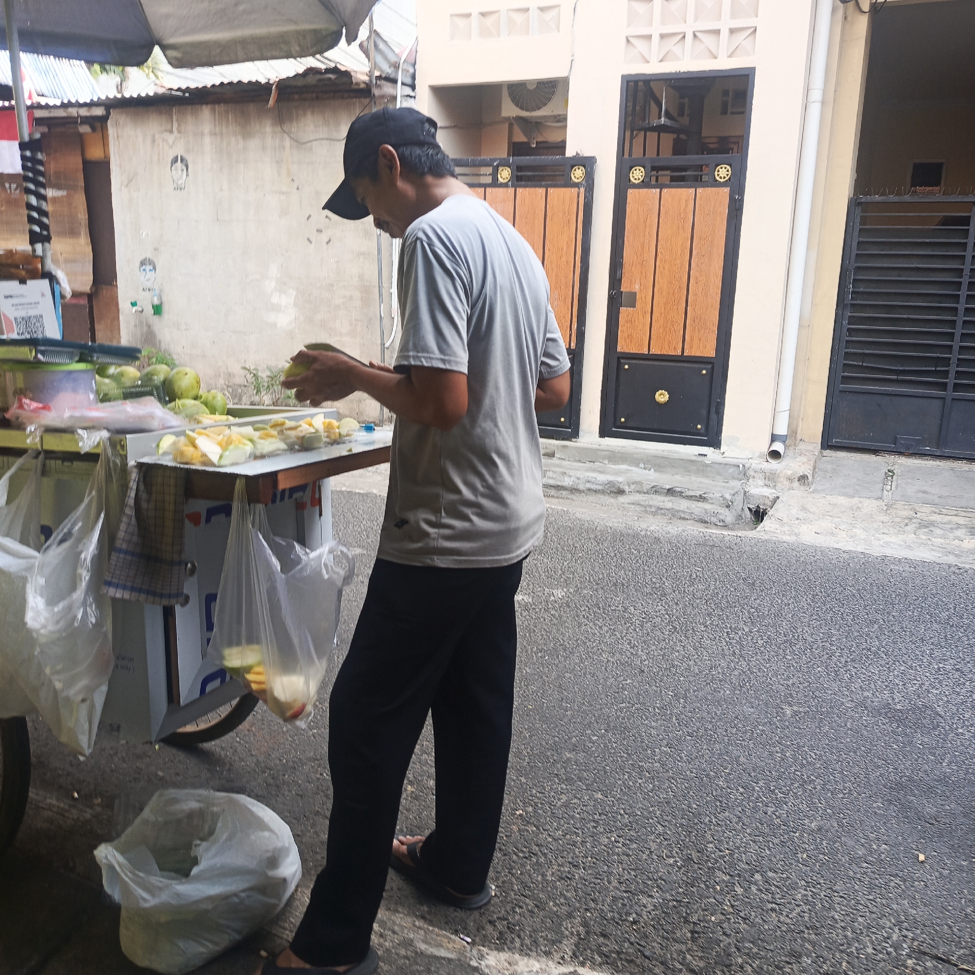 A man, wearing a gray t-shirt and a black baseball cap, stands behind a fruit cart on a paved street in front of a building. He is in the process of slicing fruit. The fruit cart is filled with various fruits, some whole and some cut, and is covered by a large umbrella. Plastic bags hang from the cart. The building in the background has wooden doors and decorative elements. The scene appears to be outdoors in an urban setting, likely South Jakarta, Indonesia, given the context of the provided information.