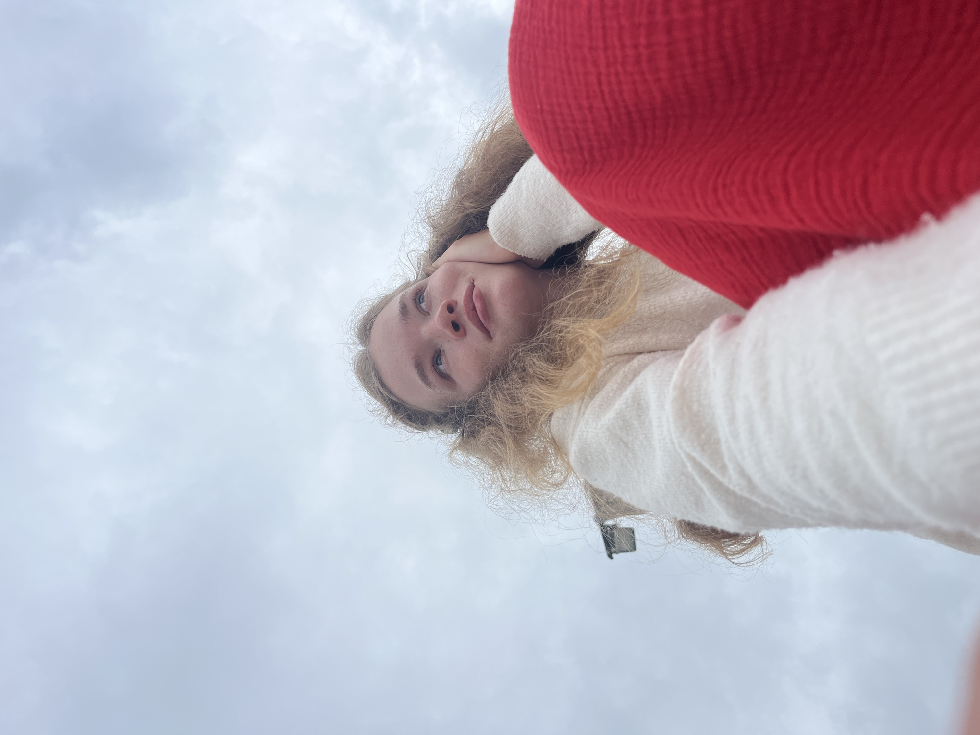 A low-angle photograph shows a young woman with blond, curly hair. She is holding her hand to her neck. She is wearing a white sweater and a red knit item. The background is a cloudy sky. The location is not identifiable as the city of Budva, Montenegro, based on the visible content.